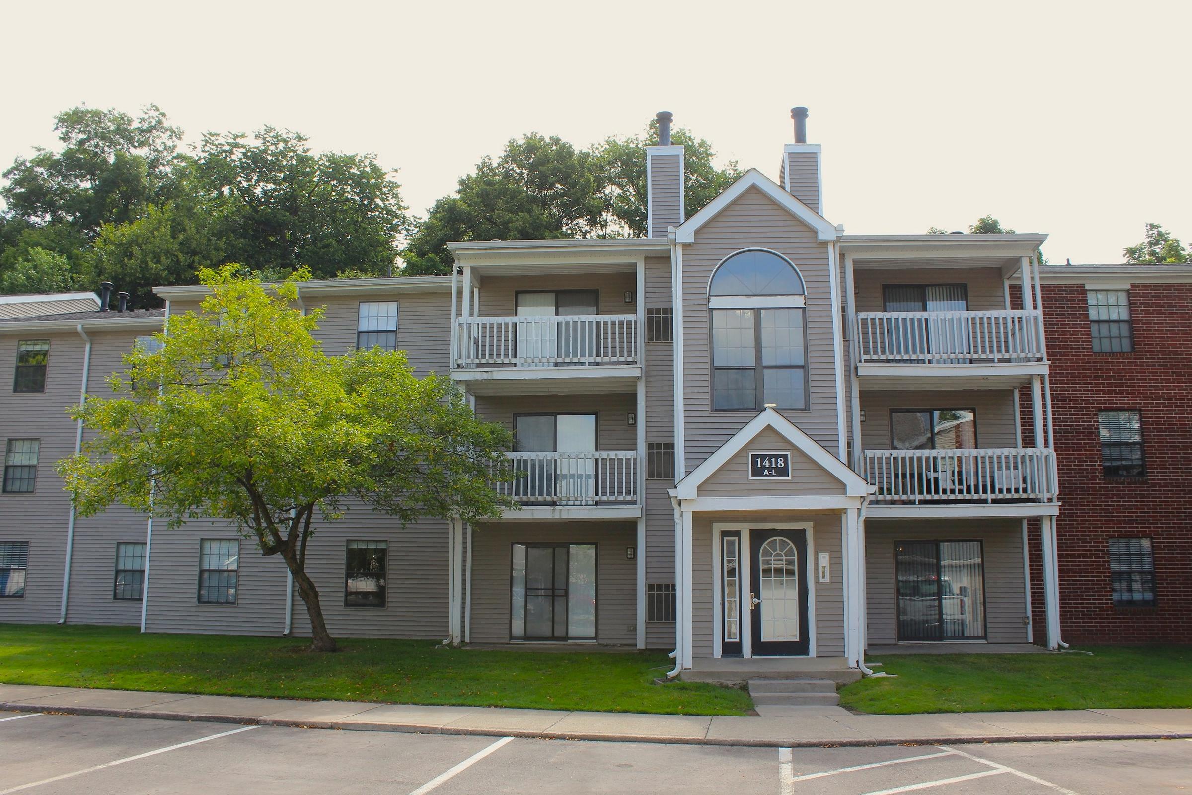 A multi-story residential building featuring a mix of gray and brick facades. It has balconies on the upper floors, a central entrance with decorative archway, and a small tree in front. The surrounding area is landscaped with grass and pavement.