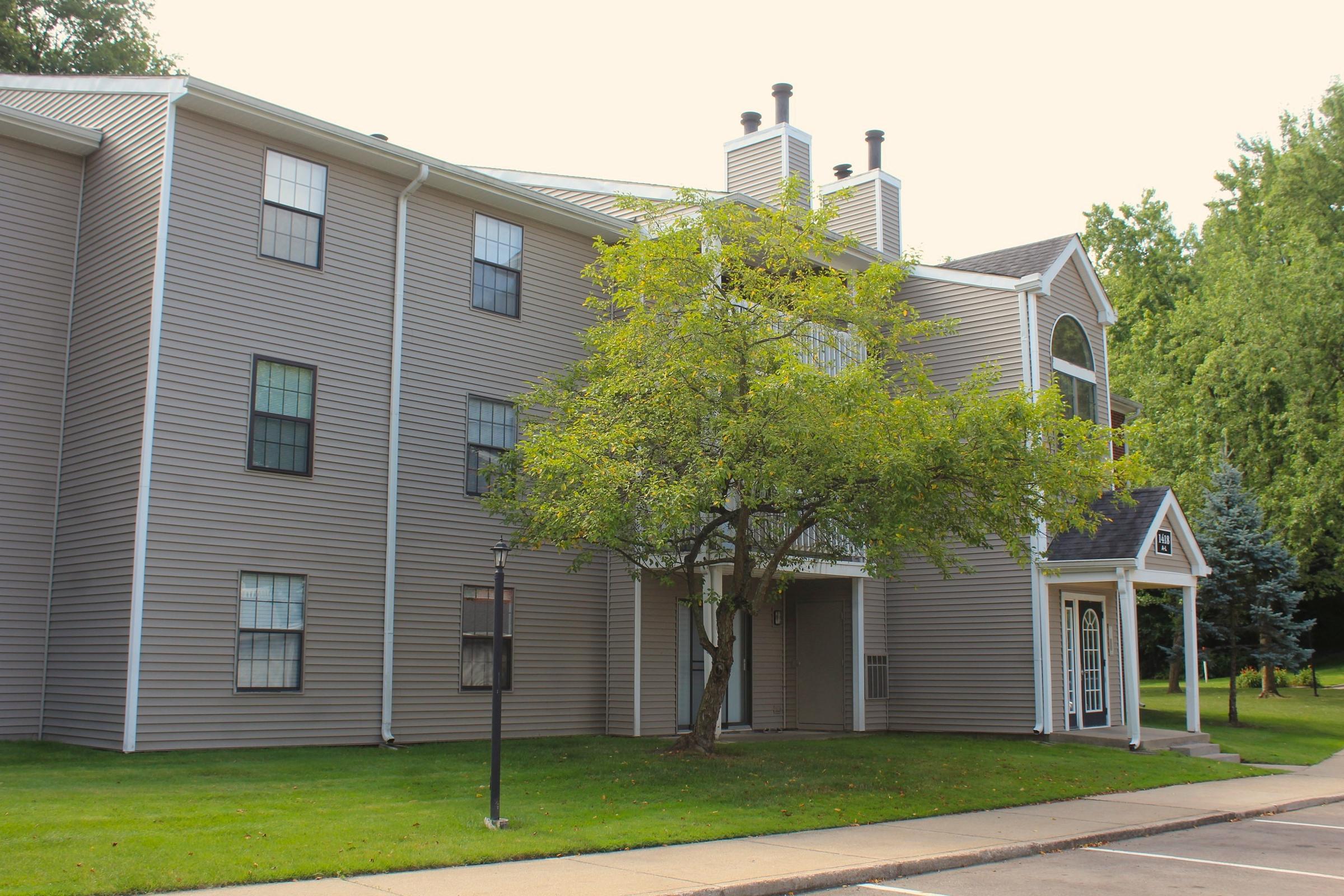 A three-story apartment building with light gray siding, featuring multiple windows. In front, there's a small tree and a grass area. The entrance includes a covered porch with a decorative roof. The surrounding landscape is well-maintained, with shrubs and trees visible in the background.