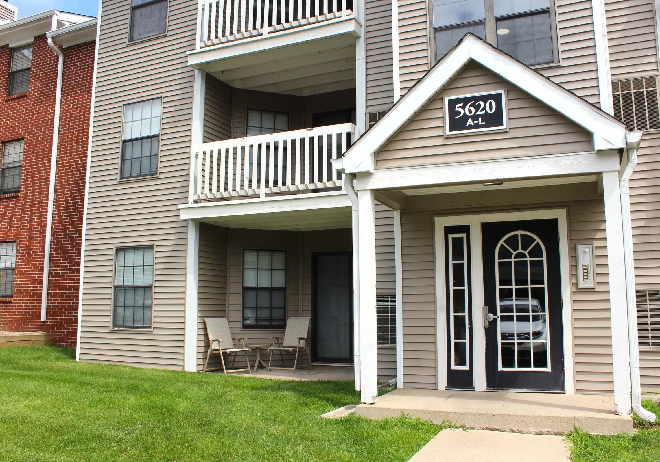Exterior view of a multi-unit apartment building with a beige facade and a black front door displaying the number "5620." Two chairs are positioned on the porch area under a small roof, with grassy landscaping and a sidewalk in front. Windows are visible on each floor.