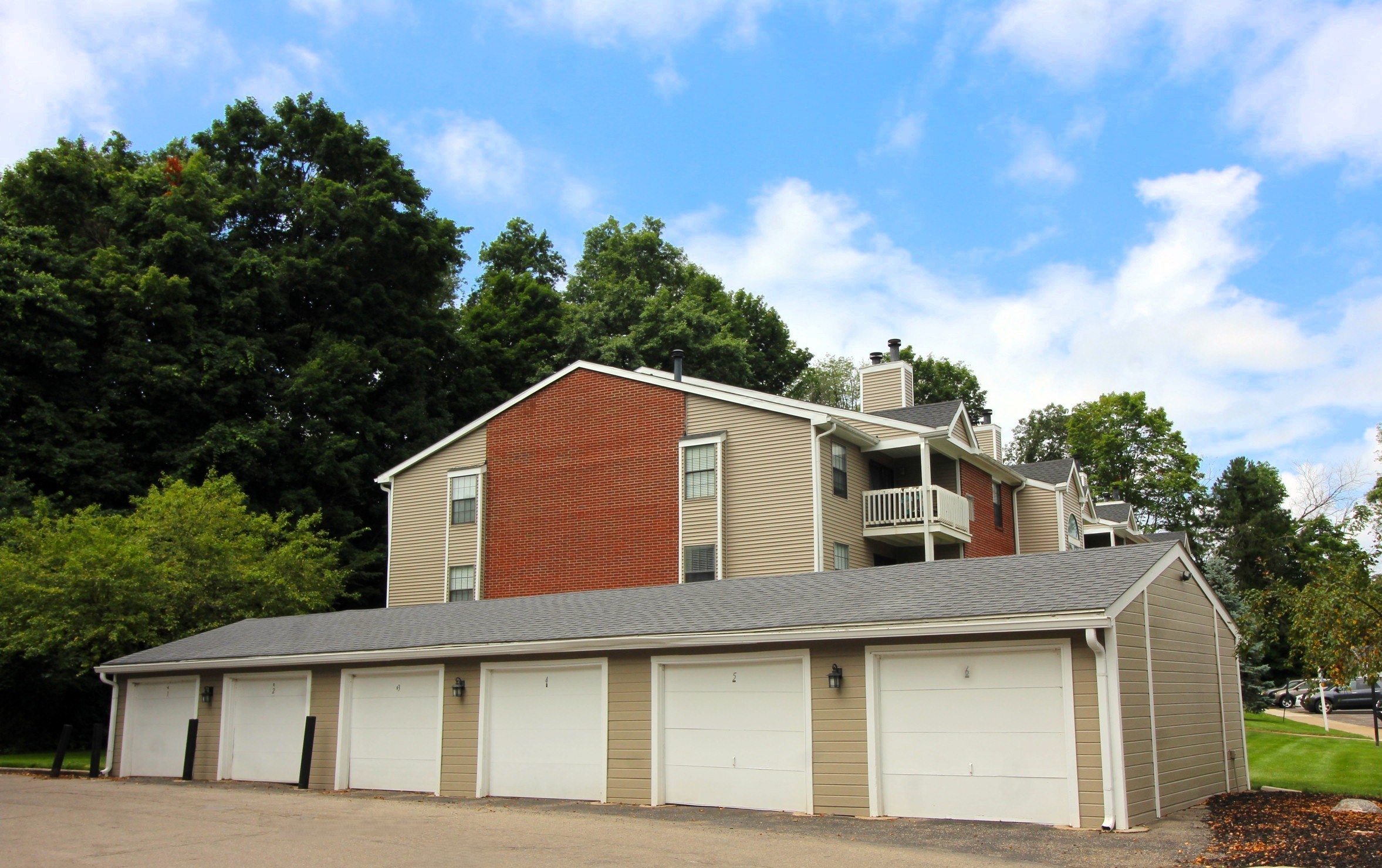 A two-story apartment building featuring a mix of brick and siding, with balconies on the upper floor. In front, there is a row of five garage doors. The setting includes green trees and a partly cloudy sky.