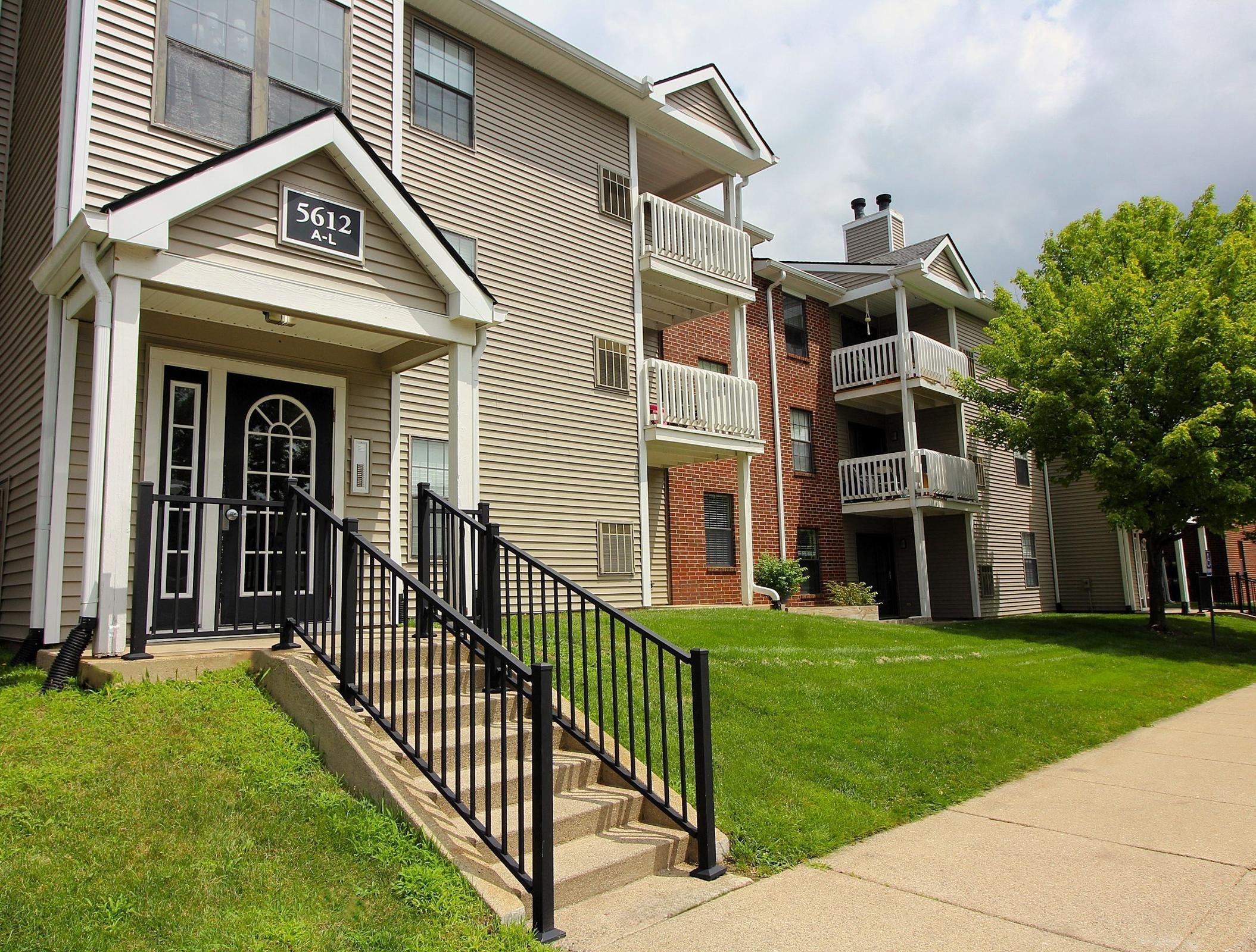 Exterior view of an apartment building featuring a staircase leading to the entrance, with balconies visible on the upper levels. Lush green lawn and trees are in the foreground. The sky is partly cloudy, creating a bright and inviting atmosphere.