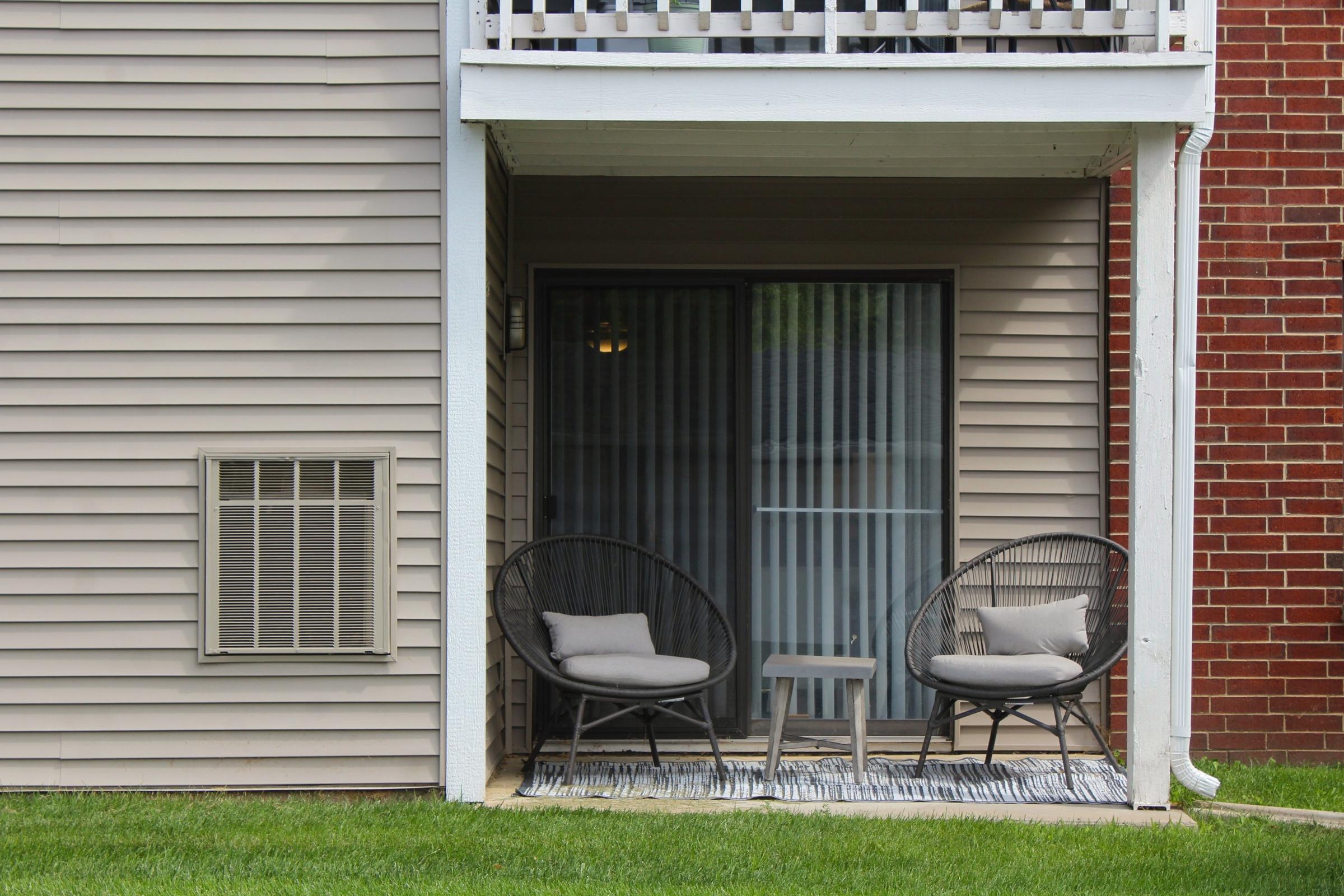 A small outdoor patio area featuring two black wicker chairs with cushions and a small table between them, situated in front of a sliding glass door, bordered by a beige building and a brick wall, with a grassy area in front.