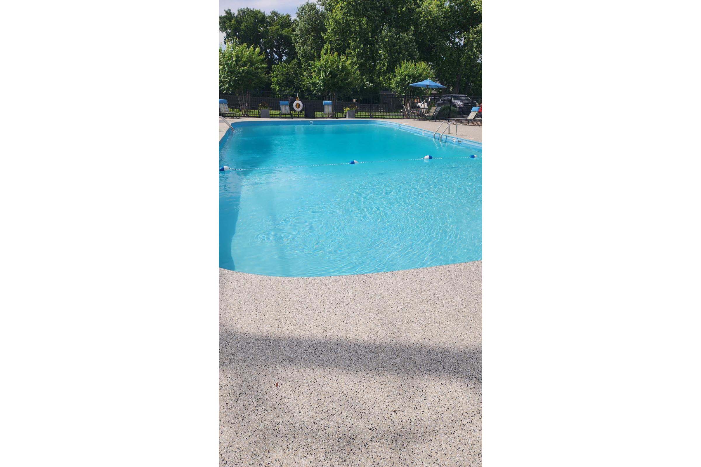 A clear blue swimming pool surrounded by a concrete deck. Lush green trees and shrubs are visible in the background, providing a serene atmosphere. Poolside chairs and umbrellas are arranged near the edge, inviting relaxation on a sunny day.