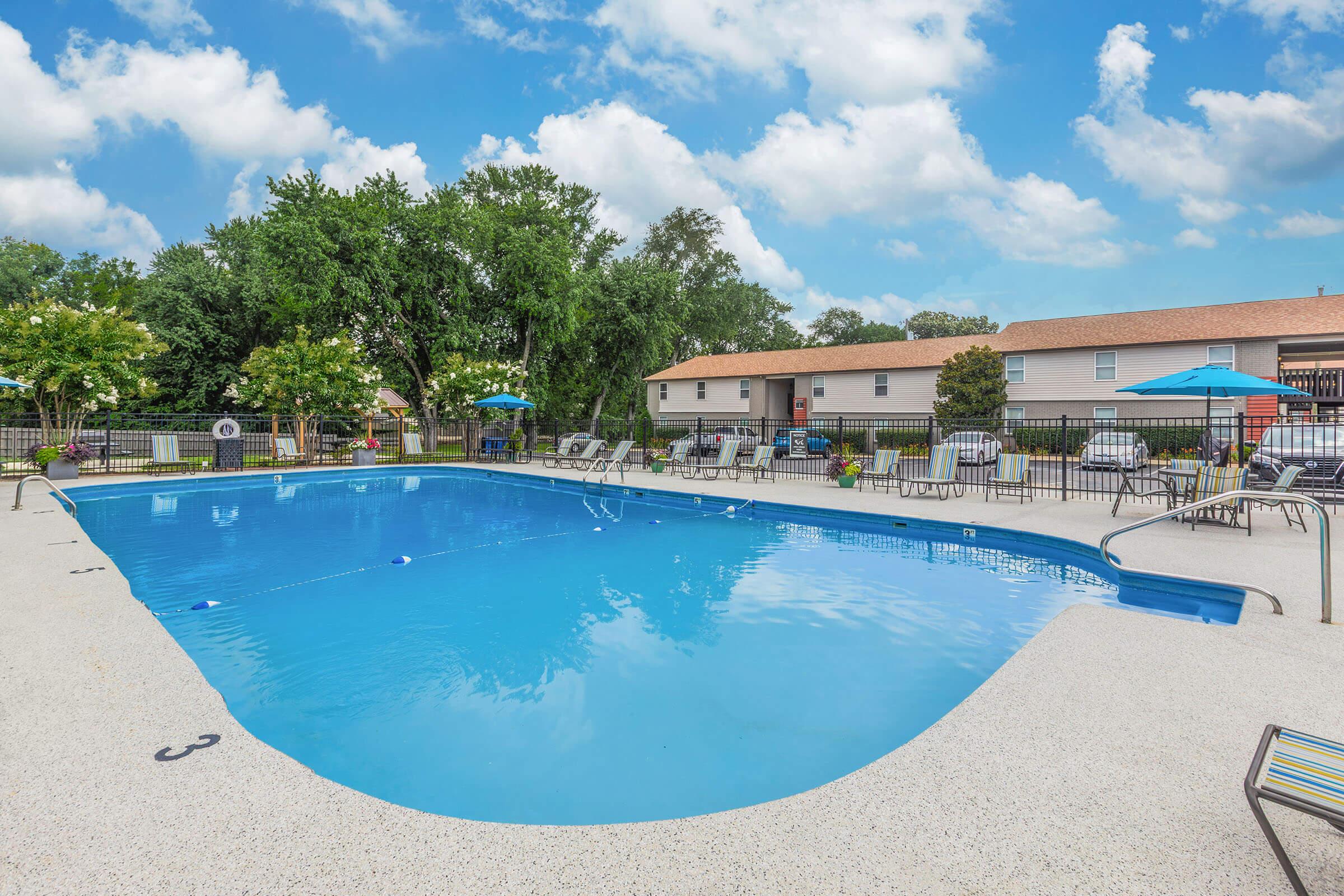 A clear blue swimming pool with lounge chairs and umbrellas around it. Green trees provide shade in the background, and a building is visible to the right. The sky is partly cloudy, creating a bright and inviting atmosphere.