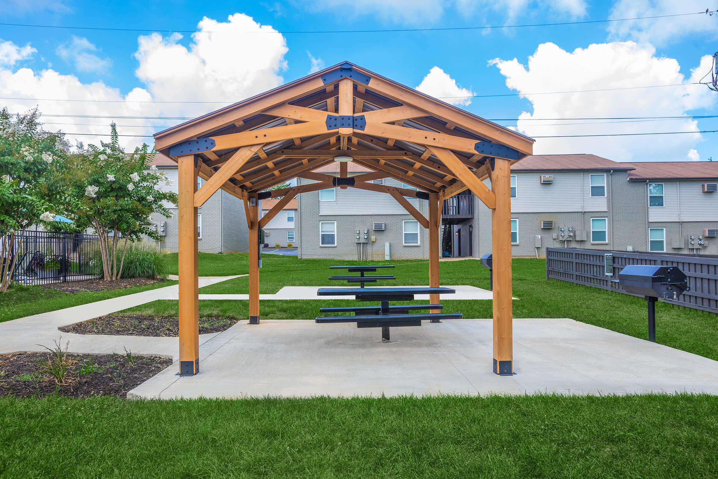 A wooden gazebo with a pitched roof and picnic tables is situated in a green park area. The gazebo is surrounded by well-maintained grass and small shrubs. In the background, there are residential buildings and a cloudy blue sky above.