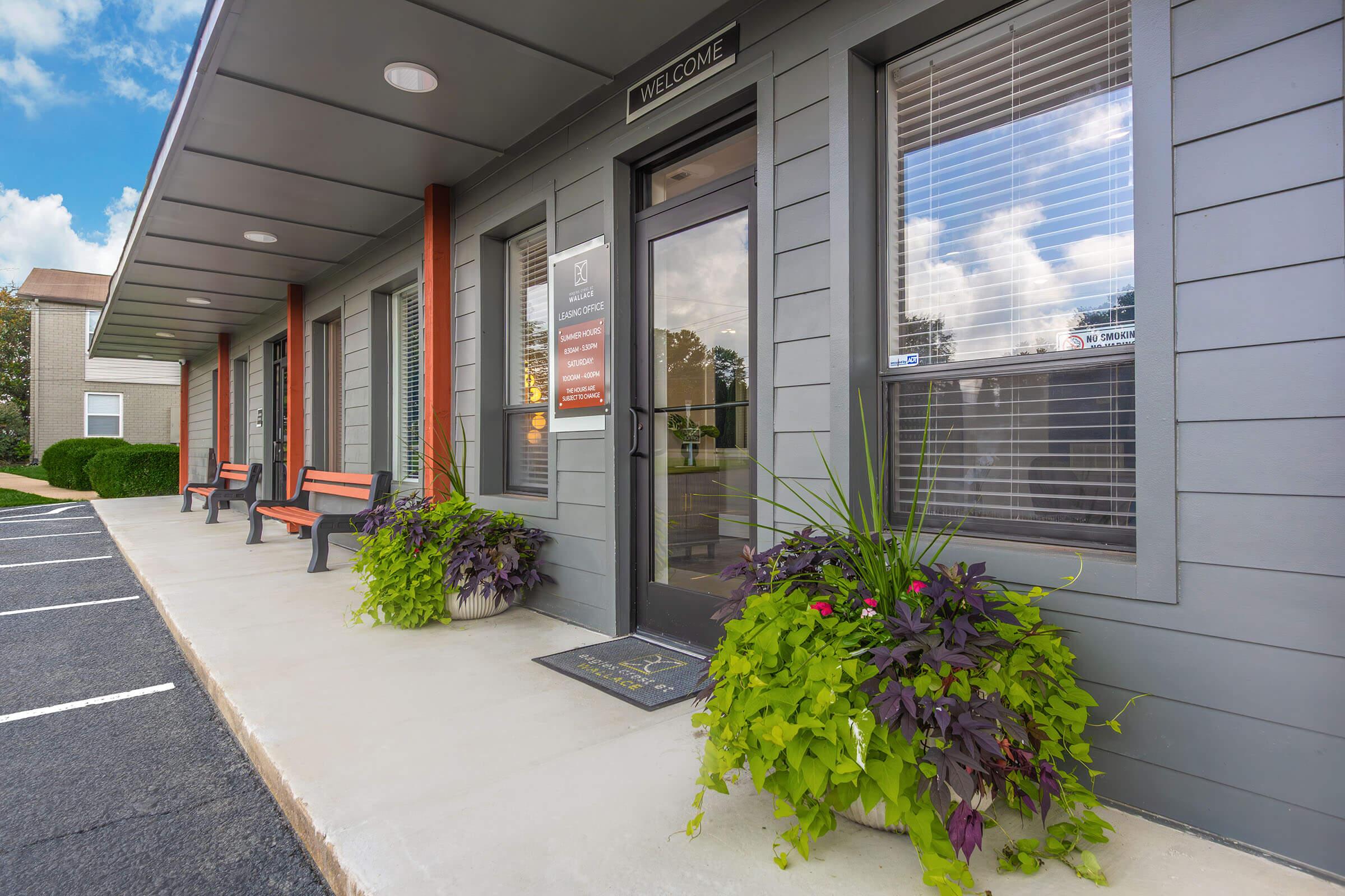 Exterior view of a welcoming building entrance featuring a "Welcome" sign, large windows with blinds, and decorative planters with vibrant plants on either side. There are wooden benches along the walkway and a clean, paved surface with marked parking spaces. The sky in the background is partly cloudy.