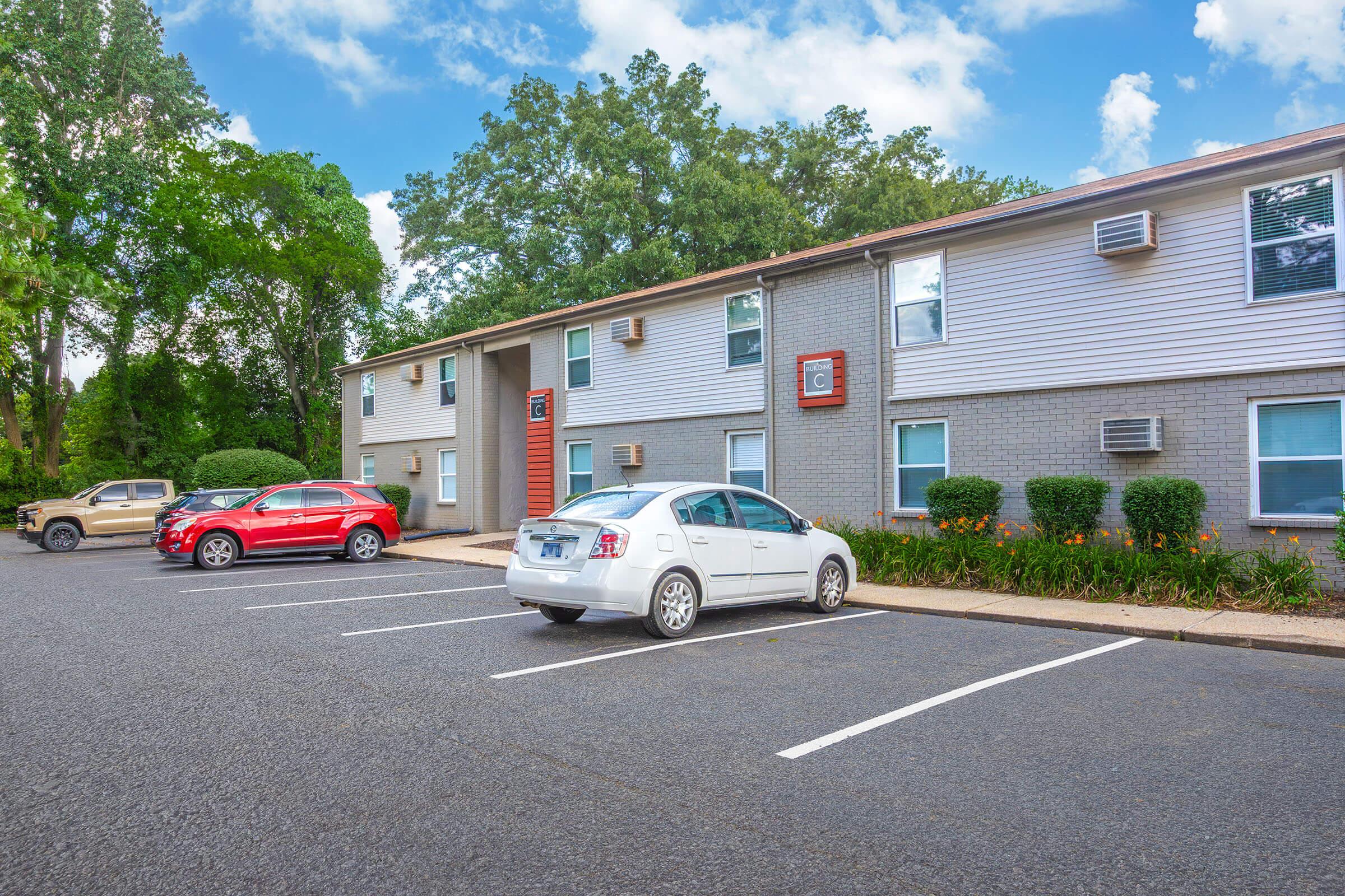 A well-maintained apartment building with a gray exterior, featuring several windows and an entrance. There are two parked cars in the foreground: a white vehicle and a red vehicle. Lush greenery and flowers are visible in front of the building, adding a touch of nature to the scene. The sky is partly cloudy.