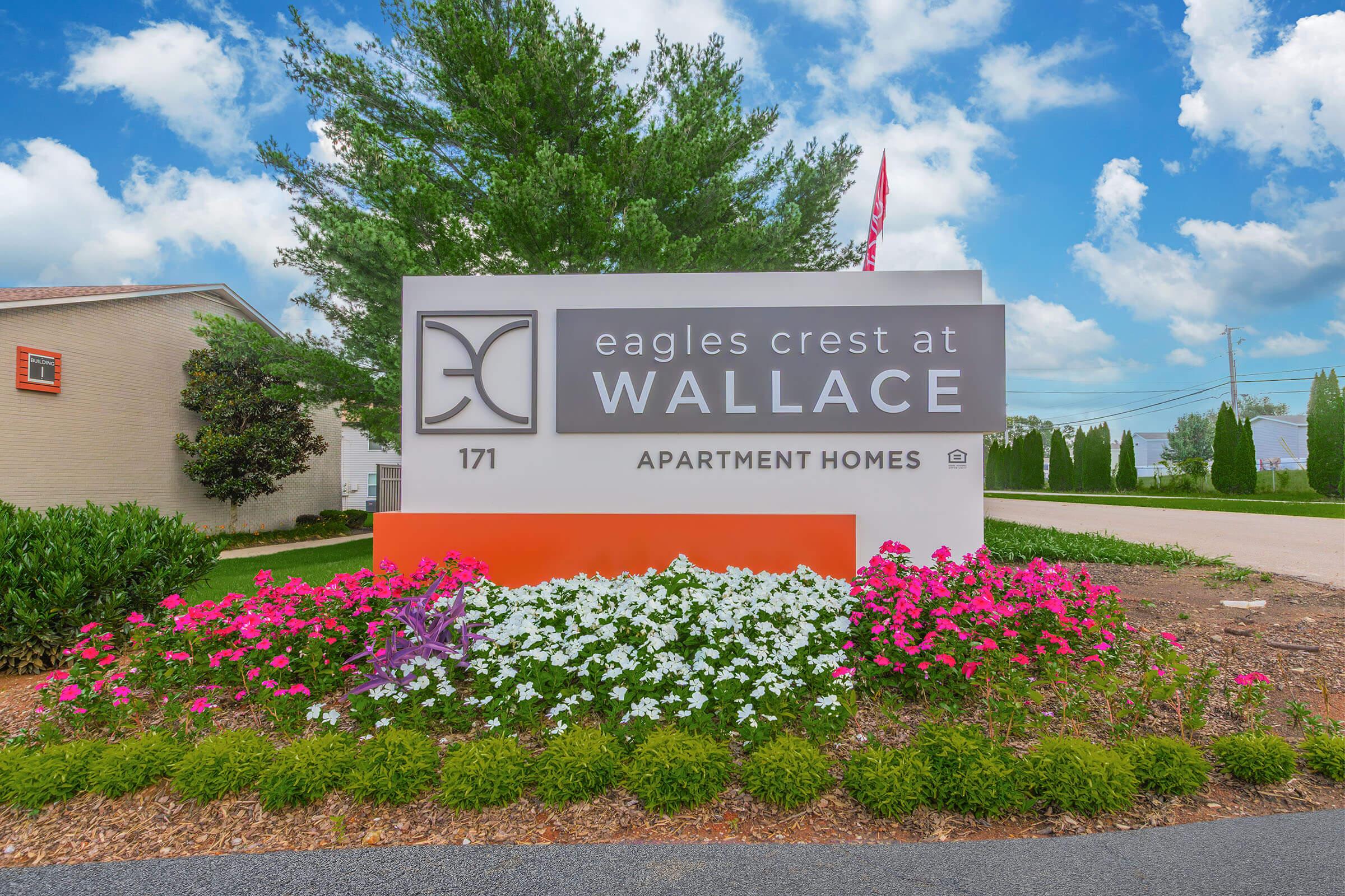 Sign for Eagles Crest at Wallace Apartment Homes, featuring the logo and address 171. The sign is surrounded by colorful flower beds and greenery, set against a bright blue sky with clouds.