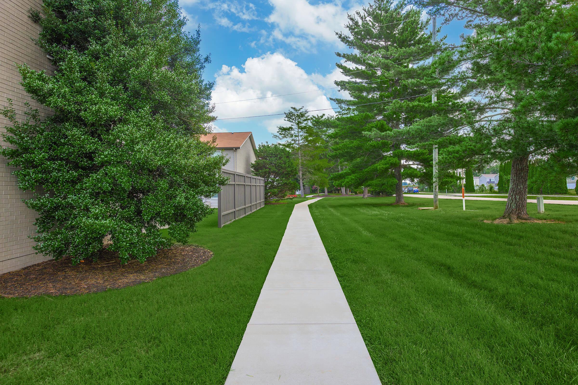 A peaceful grassy path lined with trees, leading between buildings under a blue sky with fluffy clouds. The well-maintained walkway invites leisurely strolls, surrounded by vibrant greenery and a serene atmosphere.