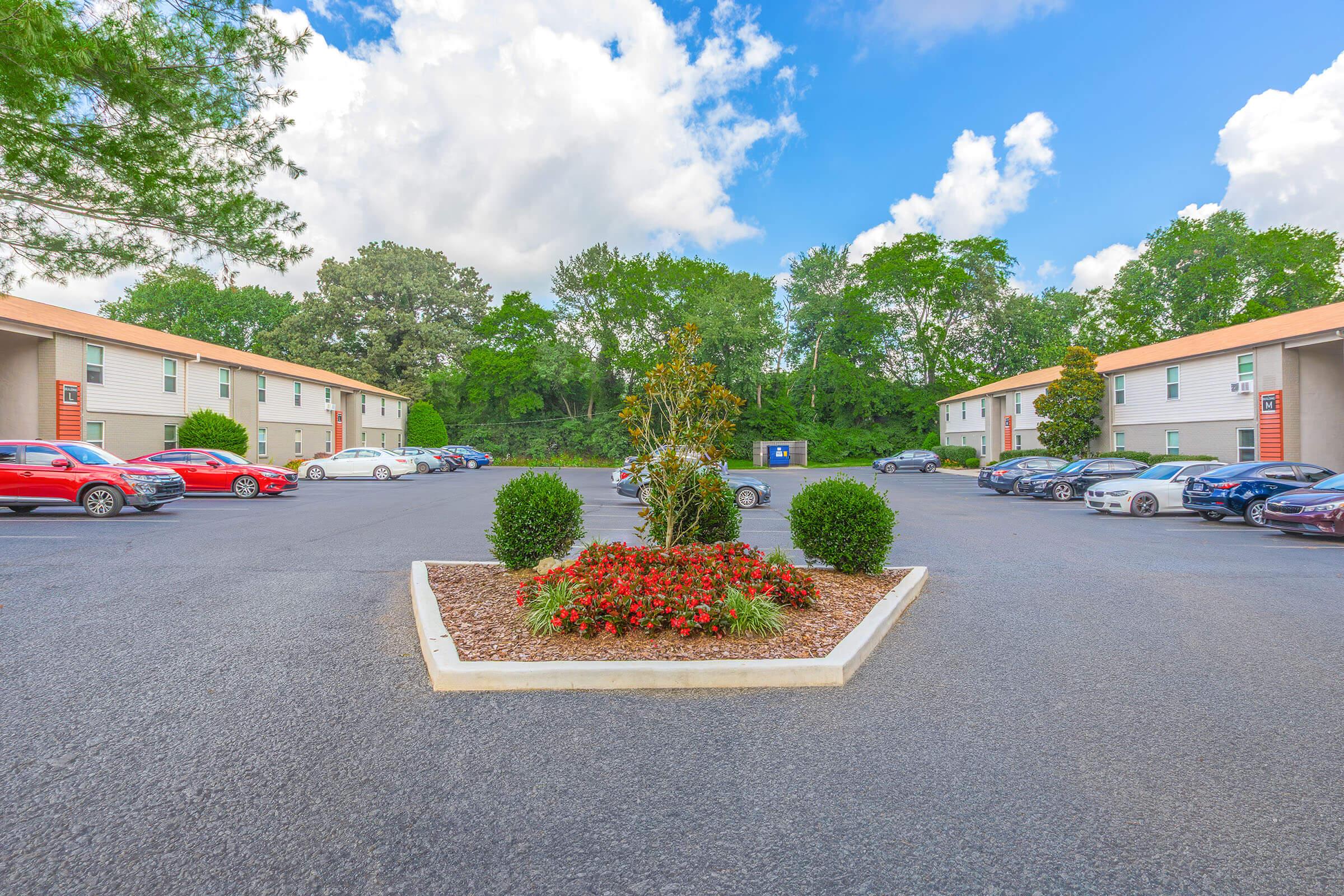 A view of a parking lot surrounded by two buildings, featuring neatly arranged bushes and flowers in the center. Several cars, including red and silver models, are parked in the lot. The background showcases trees under a partly cloudy blue sky.