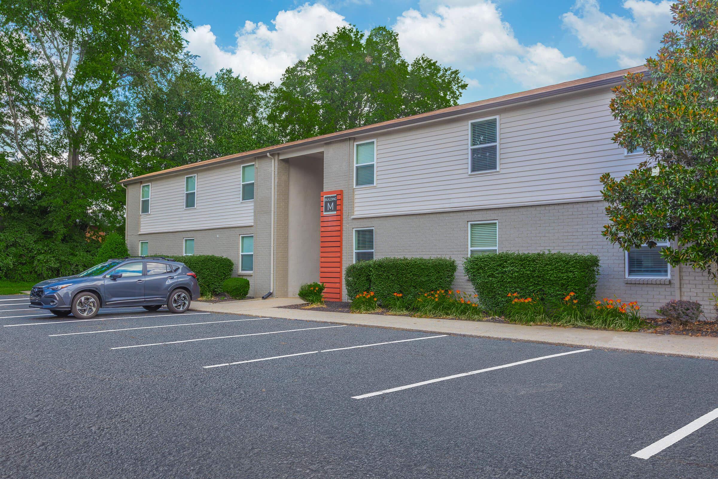 A two-story residential building with a brick and siding exterior, featuring an orange accent wall. The front has a few windows and landscaped bushes and flowers. A parked gray car is visible in the foreground, along with marked parking spaces and a tree line in the background under a partly cloudy sky.