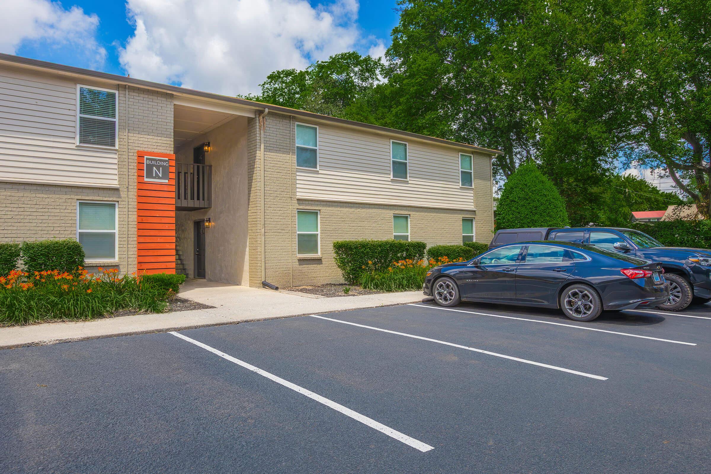 Exterior view of a modern two-story apartment building with orange and beige accents. The entrance is highlighted, surrounded by well-maintained landscaping featuring green shrubs and orange flowers. Several parked cars are in the foreground on a paved parking lot under a partly cloudy sky.
