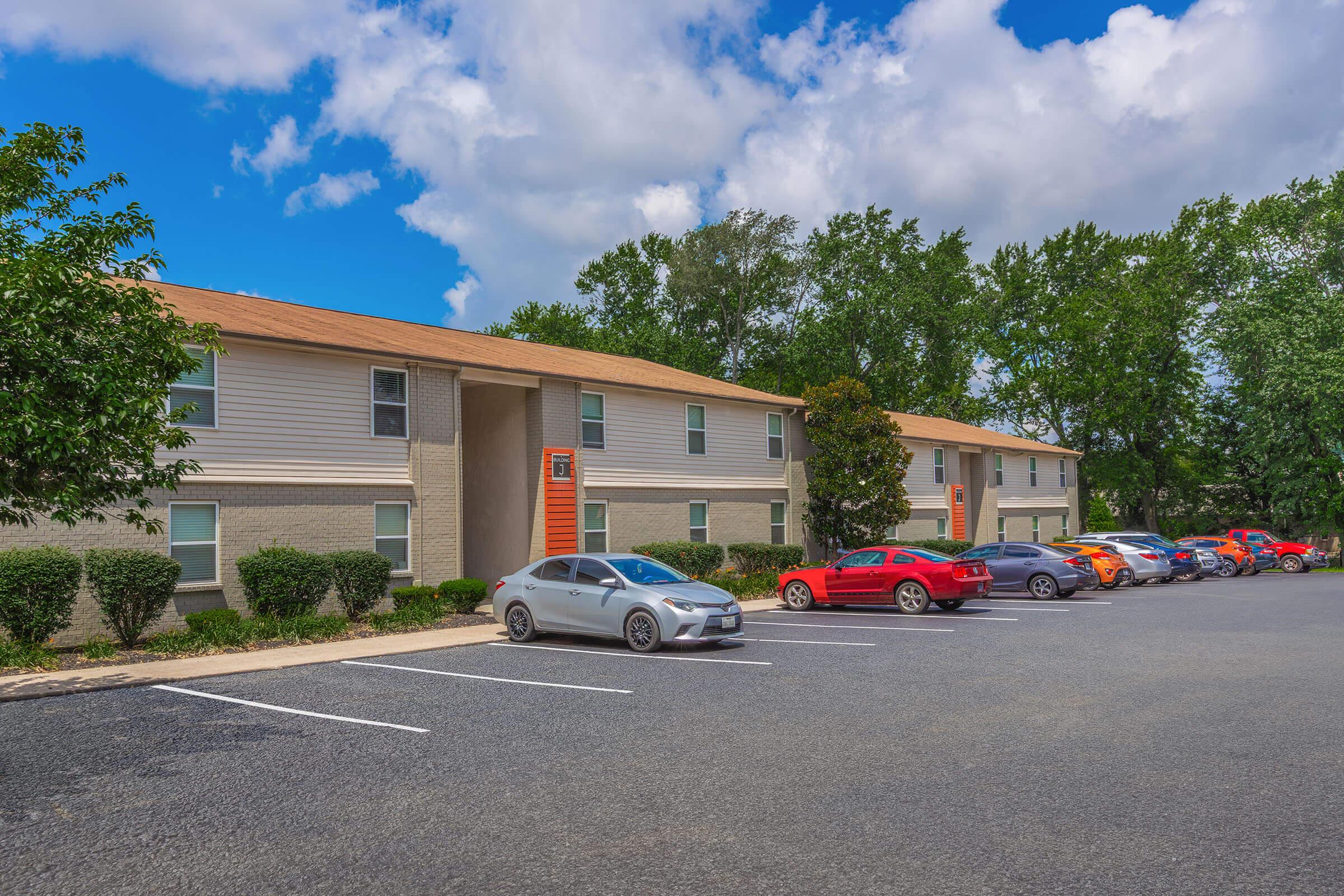 A view of a residential apartment complex featuring two-story buildings with orange and gray exteriors. Several cars are parked in the lot in front of the buildings, surrounded by green trees under a partly cloudy blue sky.