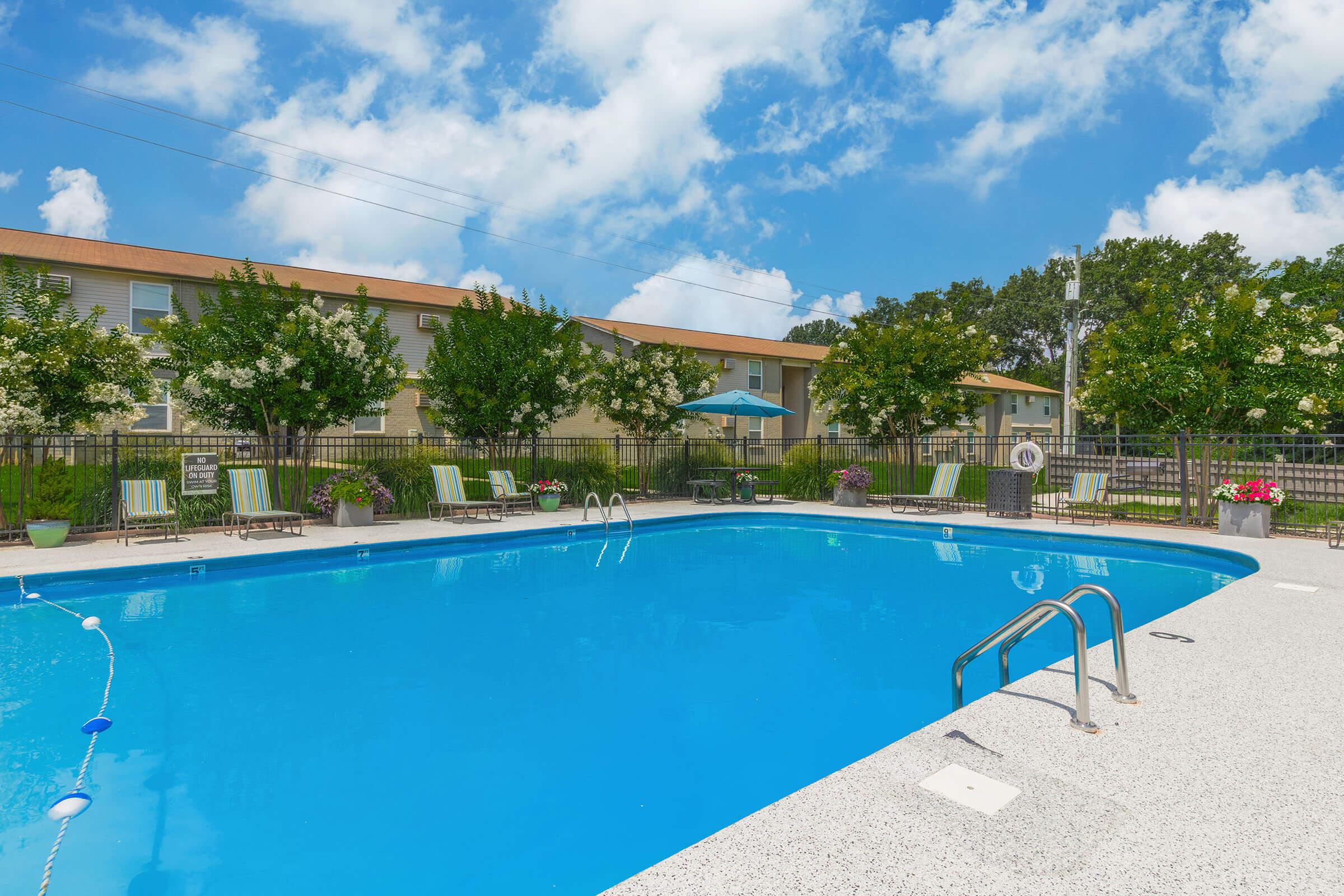 A sparkling blue swimming pool surrounded by lounge chairs and vibrant greenery. The pool area has a clean, inviting look with bright sunshine and fluffy clouds in the sky. In the background, there are buildings with well-maintained landscaping, creating a relaxing atmosphere.