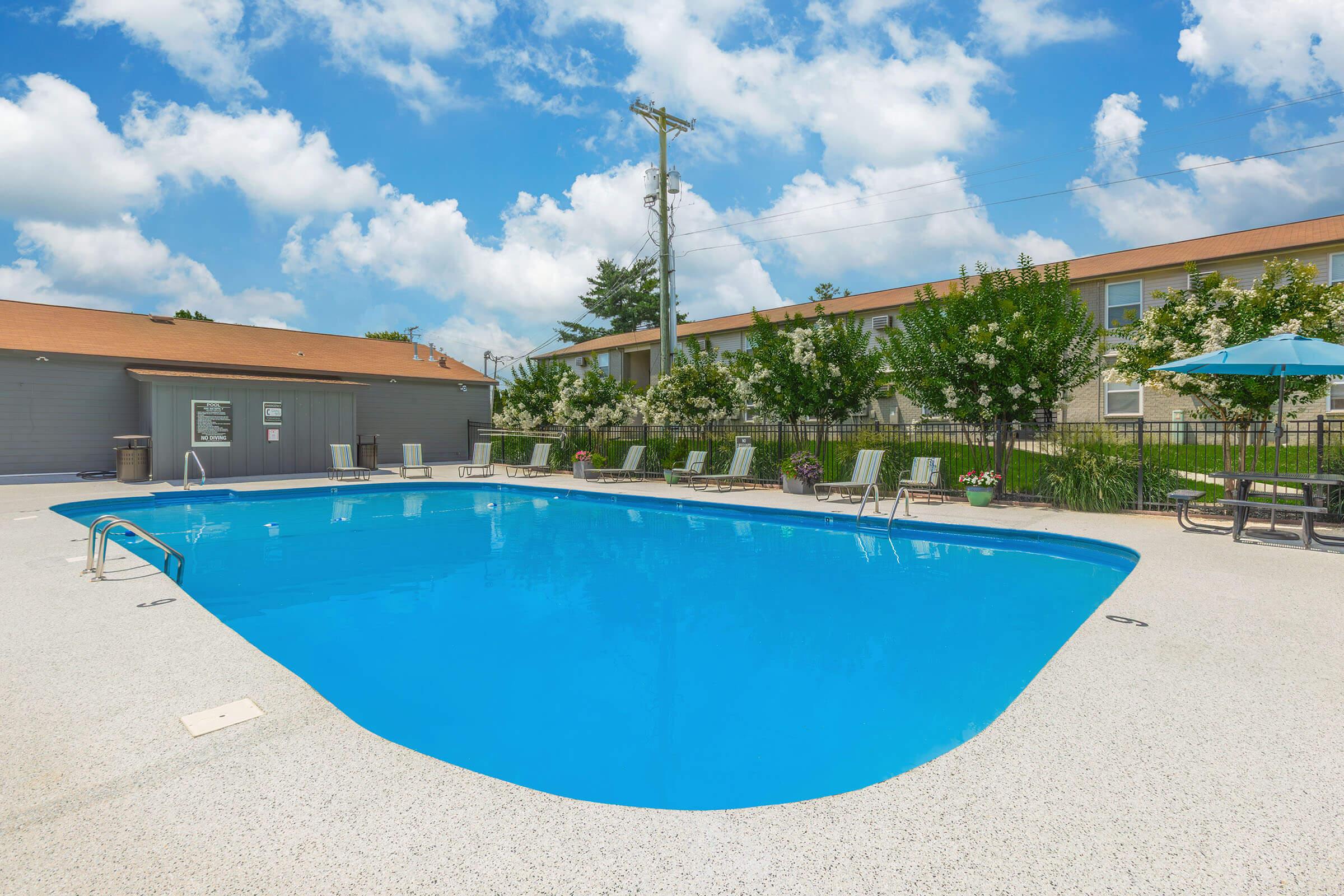 A clear blue swimming pool surrounded by lounge chairs, with a shaded seating area nearby. In the background, there are residential buildings under a bright, partly cloudy sky. The scene is well-maintained and inviting, perfect for relaxation and leisure activities.