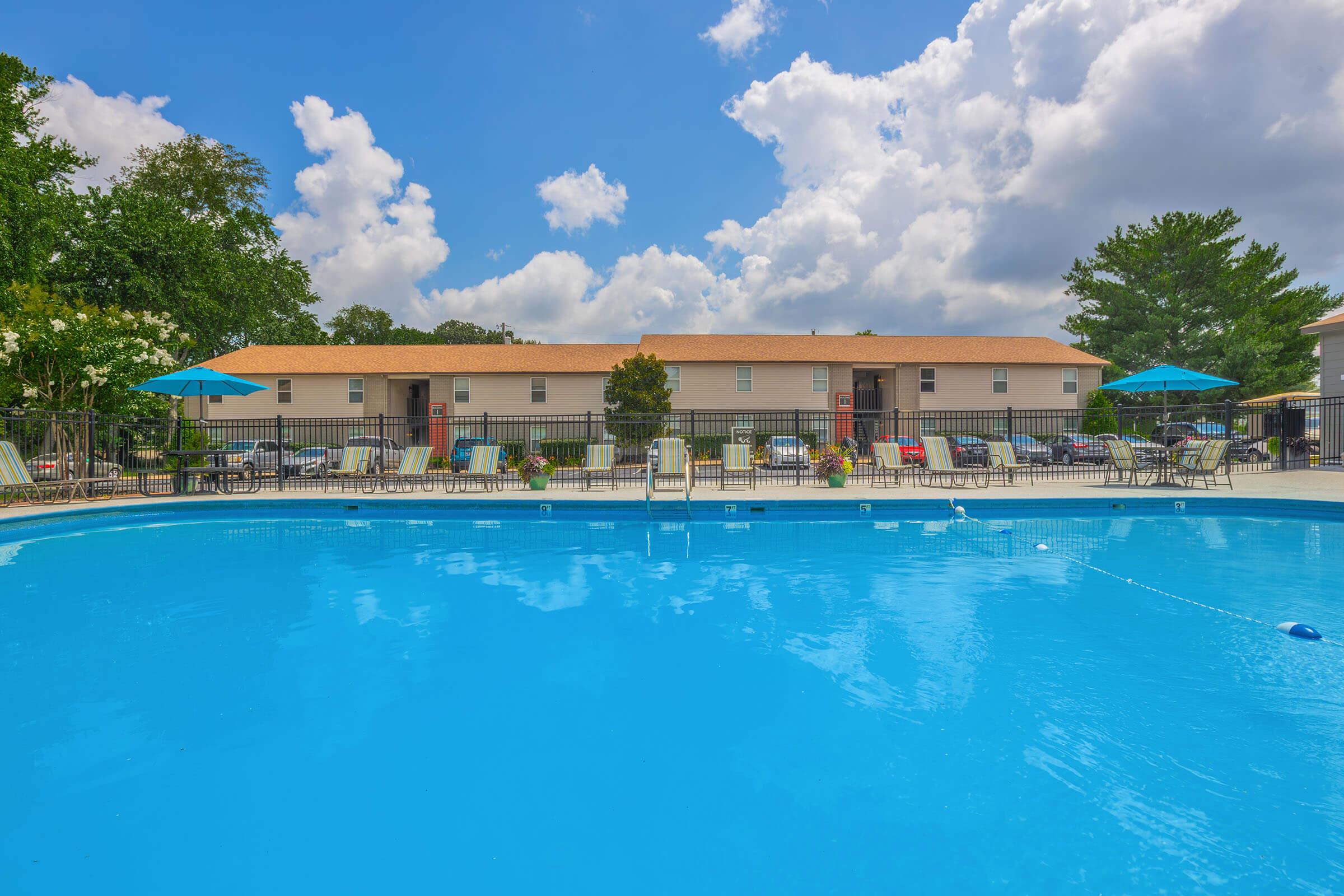 A clean, blue swimming pool surrounded by lounge chairs and tables with umbrellas. In the background, there are beige apartment buildings and lush green trees under a partly cloudy sky. The atmosphere is inviting and perfect for relaxation.