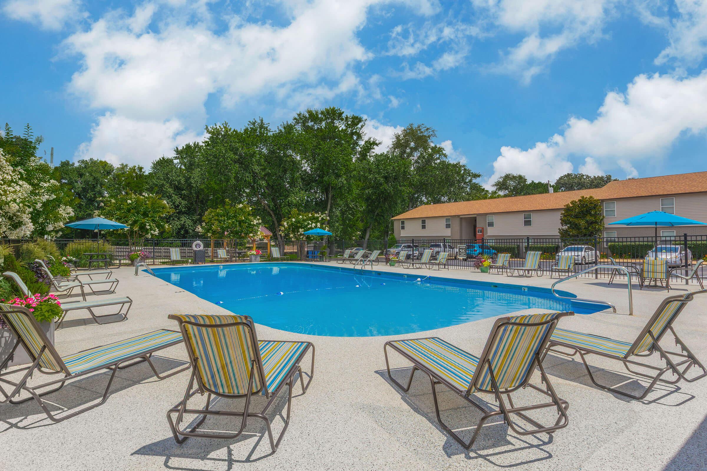 A sunny outdoor swimming pool area featuring lounge chairs with striped cushions, surrounded by greenery and bright blue umbrellas. The pool is clear and inviting, situated in a residential environment with a house in the background and a blue sky dotted with clouds.