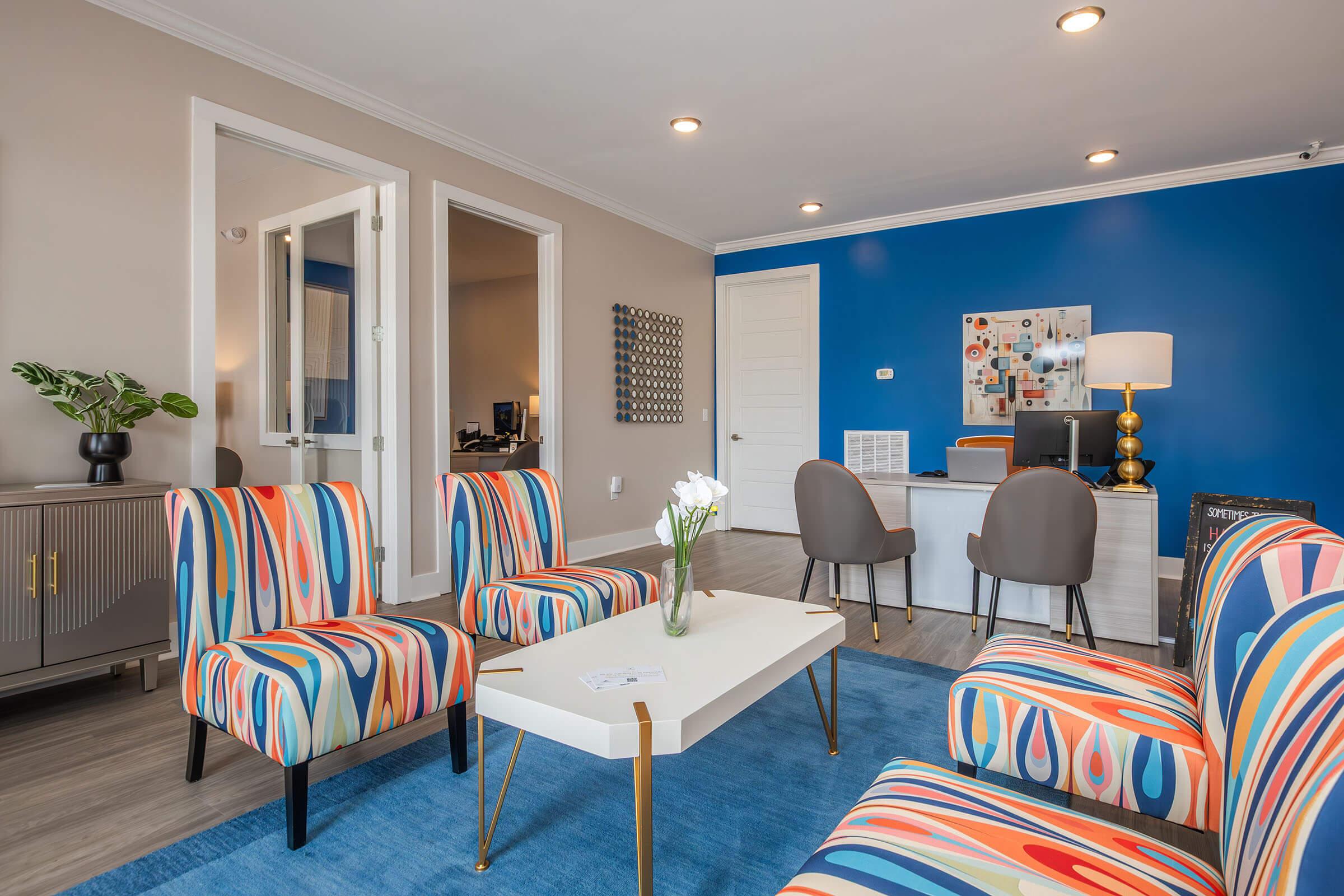 A modern office waiting area featuring colorful striped chairs and a sleek coffee table. A blue accent wall enhances the design, while a plant and a lamp add warmth. In the background, there are doors leading to other rooms, suggesting a professional environment.