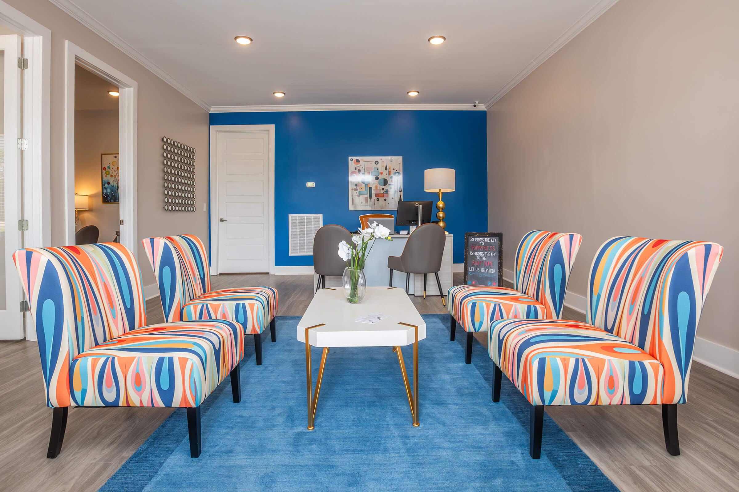 Colorful seating in a modern living space with four patterned chairs arranged around a white coffee table. A blue area rug lies beneath, while a blue accent wall features artwork. Additional seating and a lamp are visible in the background, creating a vibrant and inviting atmosphere.