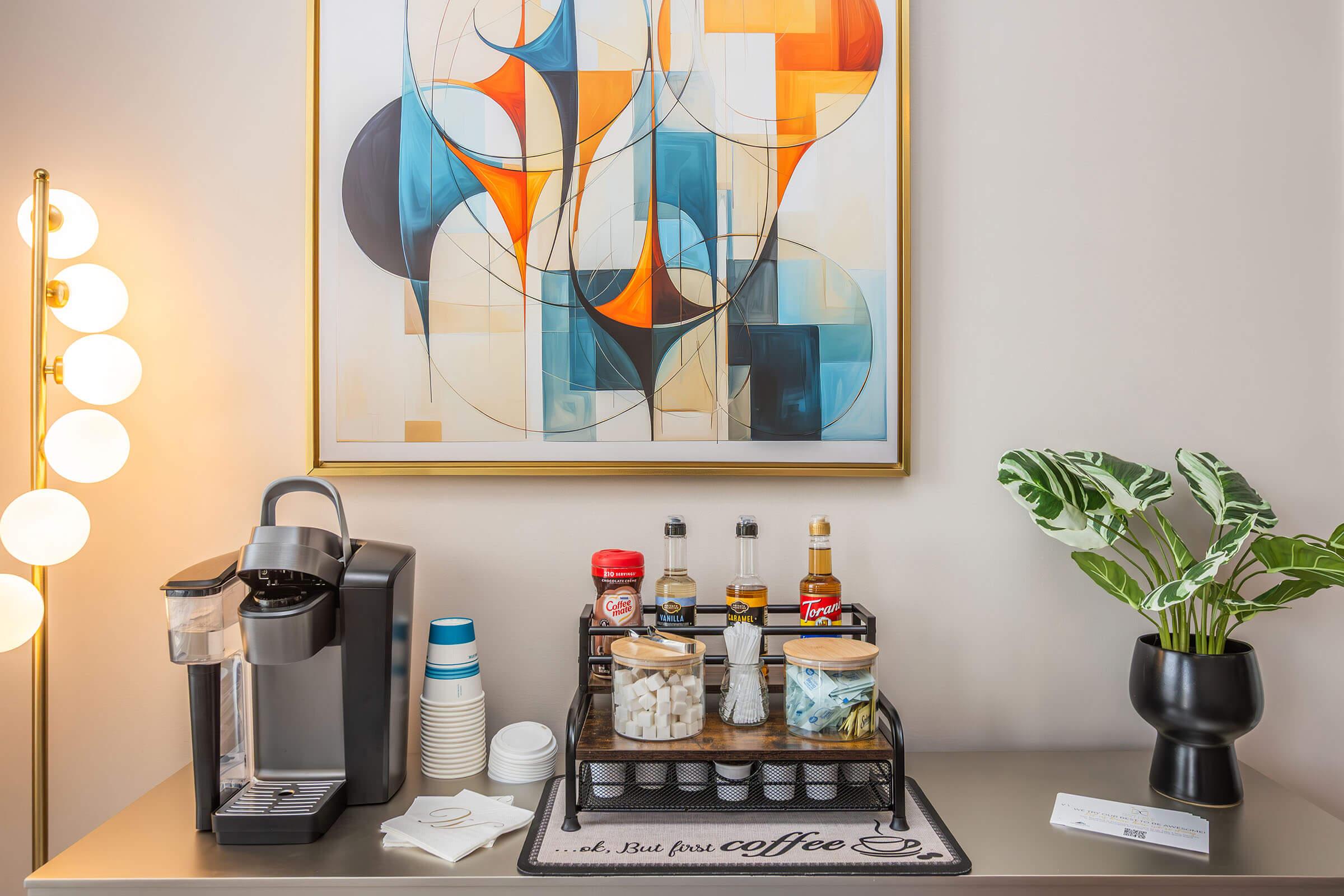 A modern coffee station featuring a coffee maker, various condiments in jars, disposable cups, and a decorative area rug with "But first, coffee" printed on it. A vibrant abstract painting hangs above the station, and a potted plant adds a touch of greenery to the setup. Soft lighting enhances the aesthetic.