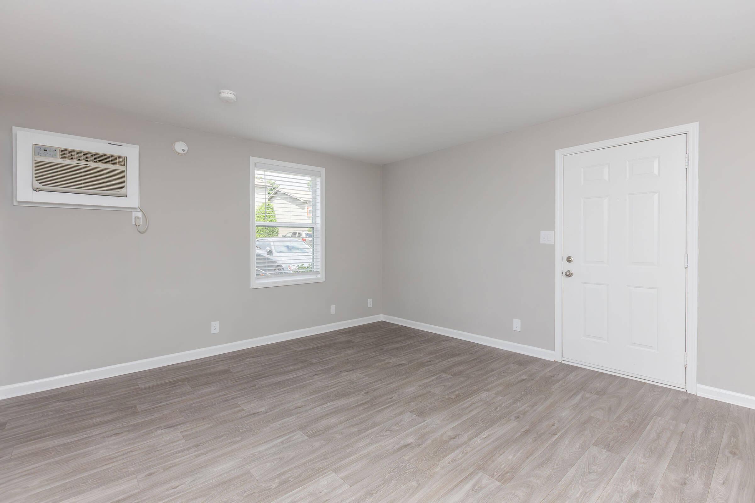 A spacious, empty room with light gray walls and a wooden floor. There is an air conditioning unit on the wall, a window with white blinds, and a closed white door visible. The room has a clean and modern appearance, suitable for various uses.