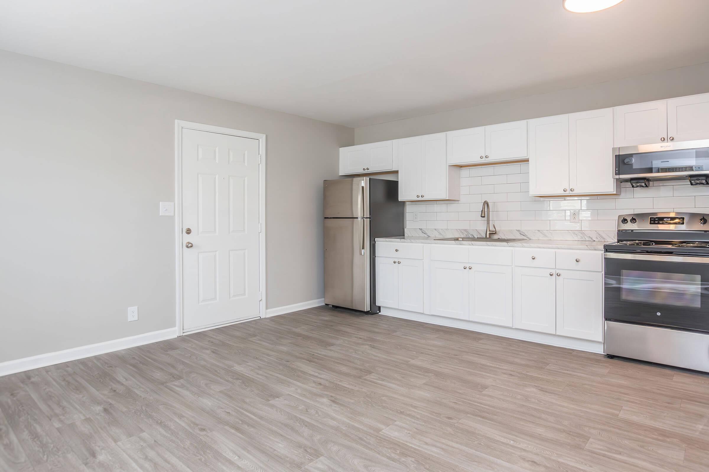 Modern kitchen interior with light gray walls, laminate flooring, and white cabinetry. Features include a stainless steel refrigerator, a stove with an oven, and a sink with a minimalist faucet. A door leads to another room, and natural light fills the space.