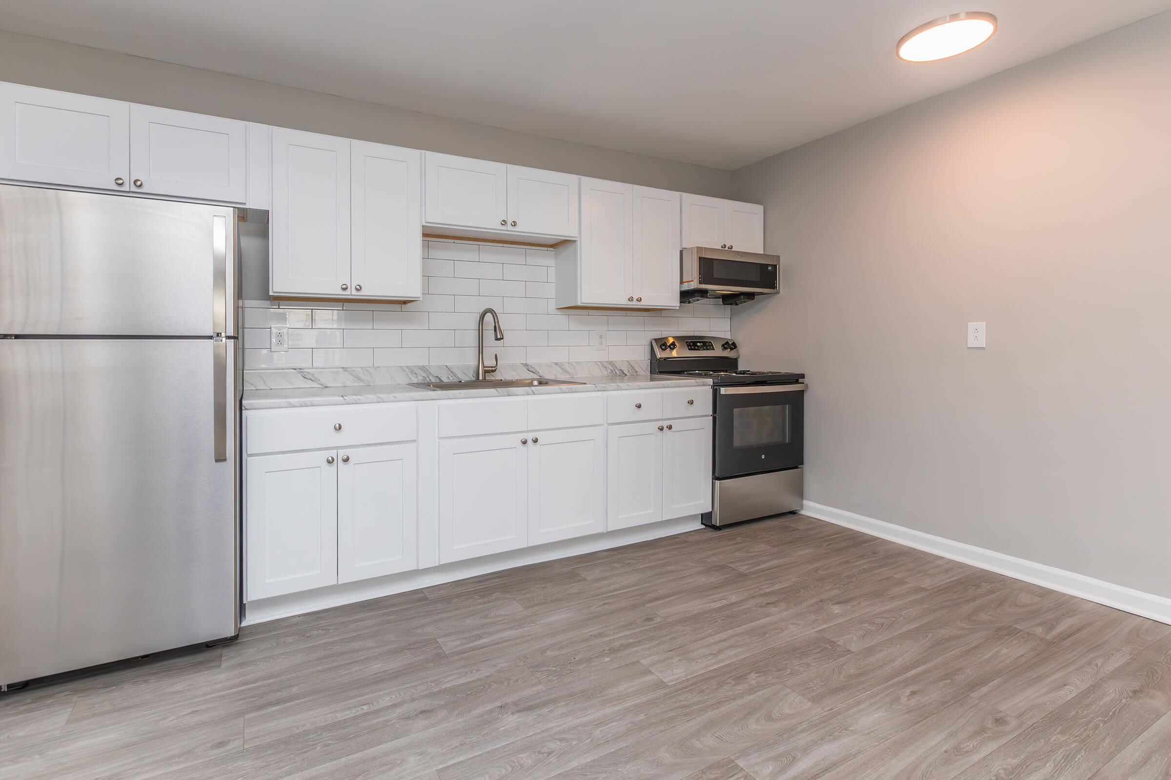 A modern kitchen with white cabinets, a stainless steel refrigerator, an oven, and a microwave. The countertop features a marble pattern, and there's a sink installed beneath a window. The floor is made of light-colored wood laminate, and the walls are painted in a neutral shade.
