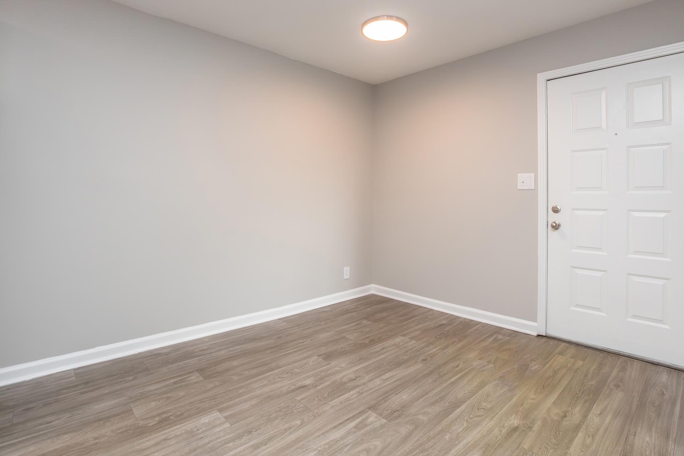 Empty room with light gray walls and a laminate floor. A white door with a doorknob is visible to the right side, and a ceiling light is on. The space has no furniture or decorations, creating a minimalist and clean aesthetic.