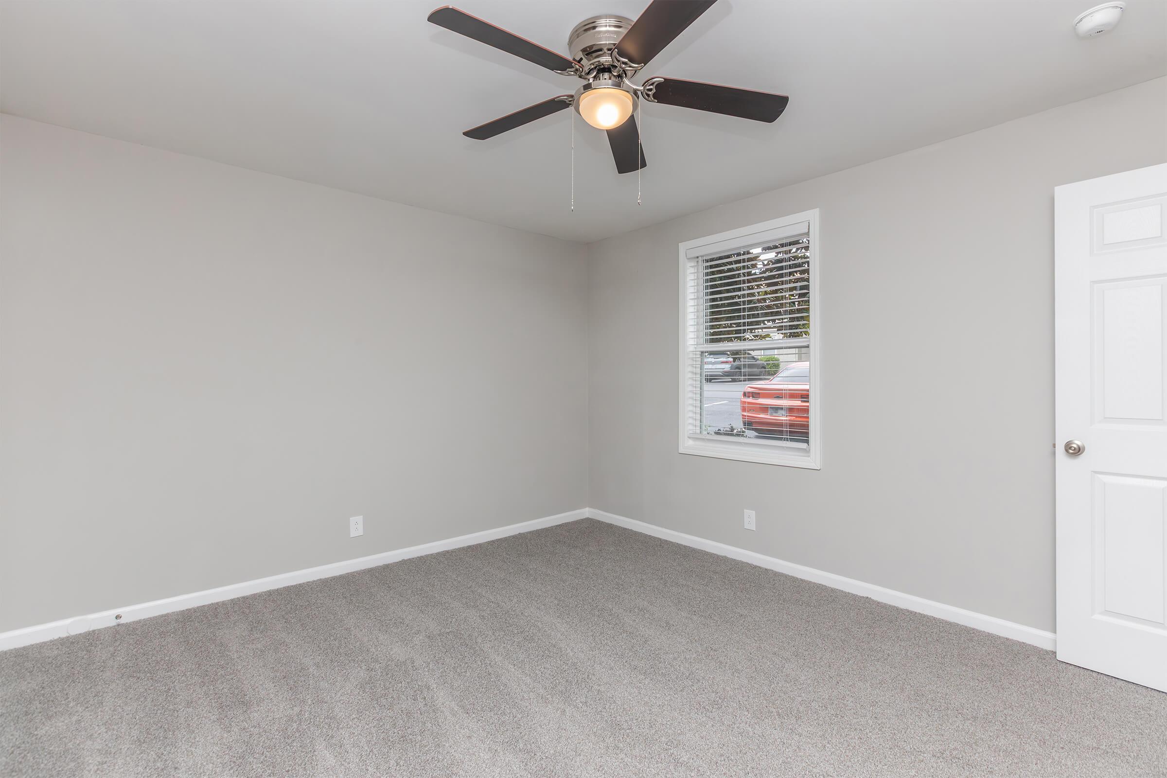 Empty room with light gray walls and new carpet. Features a ceiling fan with light, a single window allowing natural light, and a white door. The room has a minimalist design and appears well-lit, creating an open and airy atmosphere.