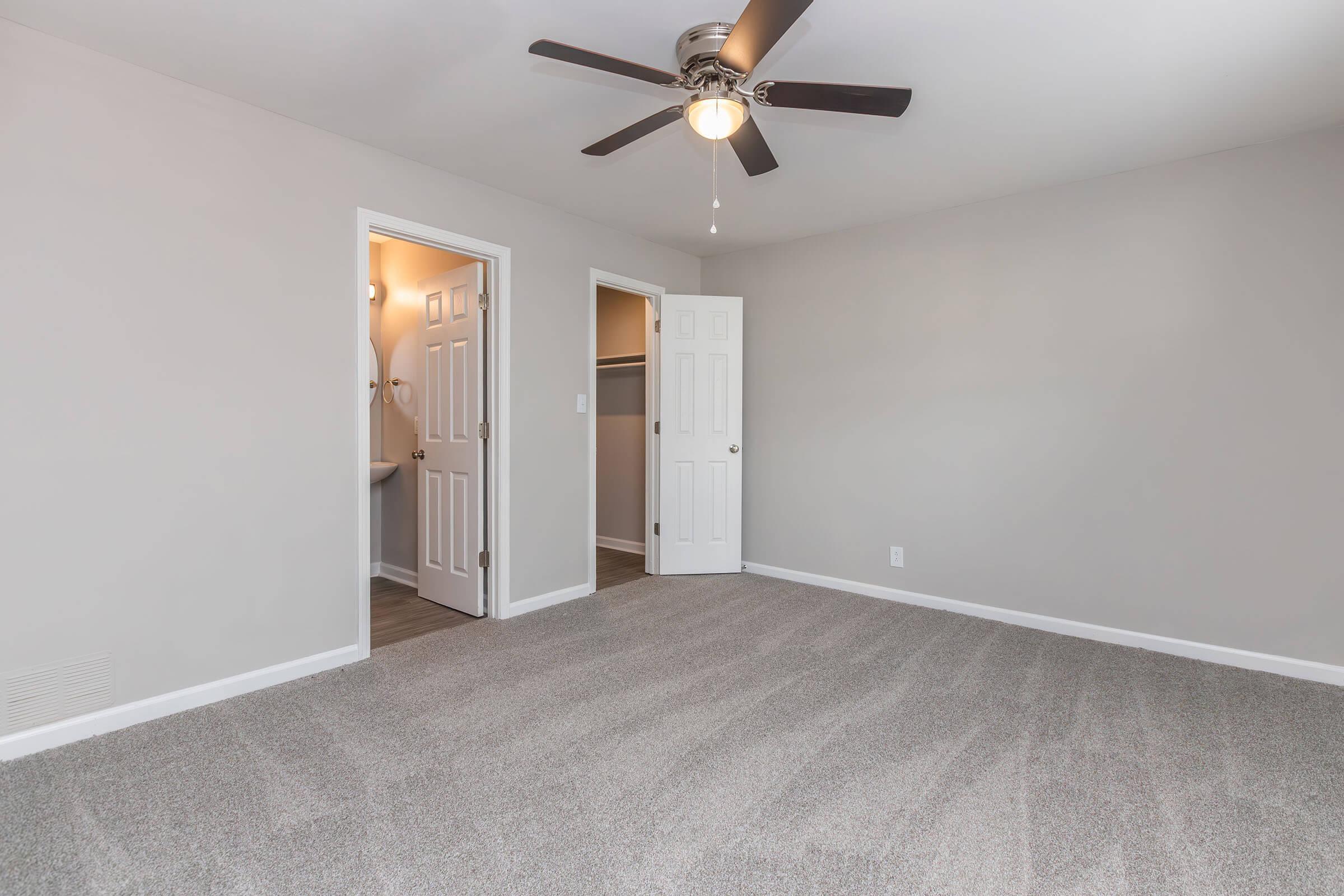 A well-lit bedroom featuring light gray walls and new carpet. The room includes a ceiling fan and two open doors leading to closets or a bathroom. Natural light invites a cozy feel, making it suitable for relaxation or sleeping.