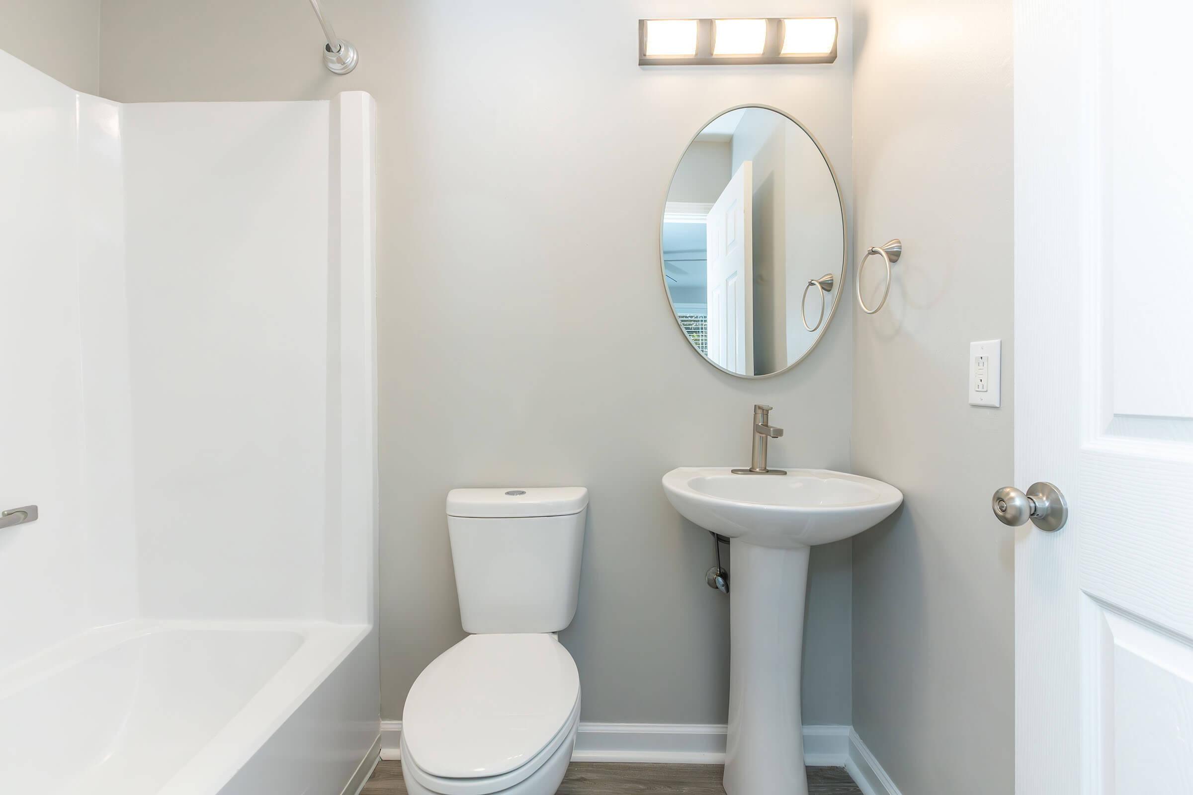 A modern bathroom featuring a white bathtub and shower combination, a toilet, a pedestal sink, and a round mirror. The walls are painted light gray, and there is a light fixture above the mirror. The floor has a wooden appearance, contributing to a clean and minimalist aesthetic.