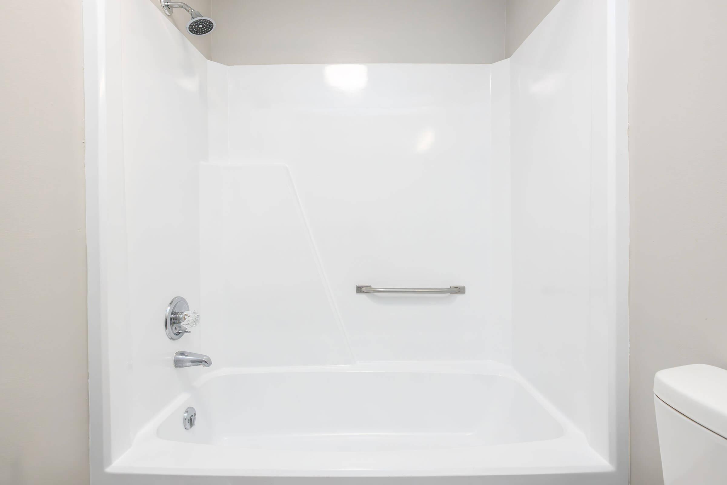 A clean, modern bathroom shower area featuring a white tub and shower unit. The shower has a sleek chrome handle and a showerhead mounted on the wall. The walls are a neutral color, creating a bright and airy ambiance. A toilet can be seen partially on the right side of the image.