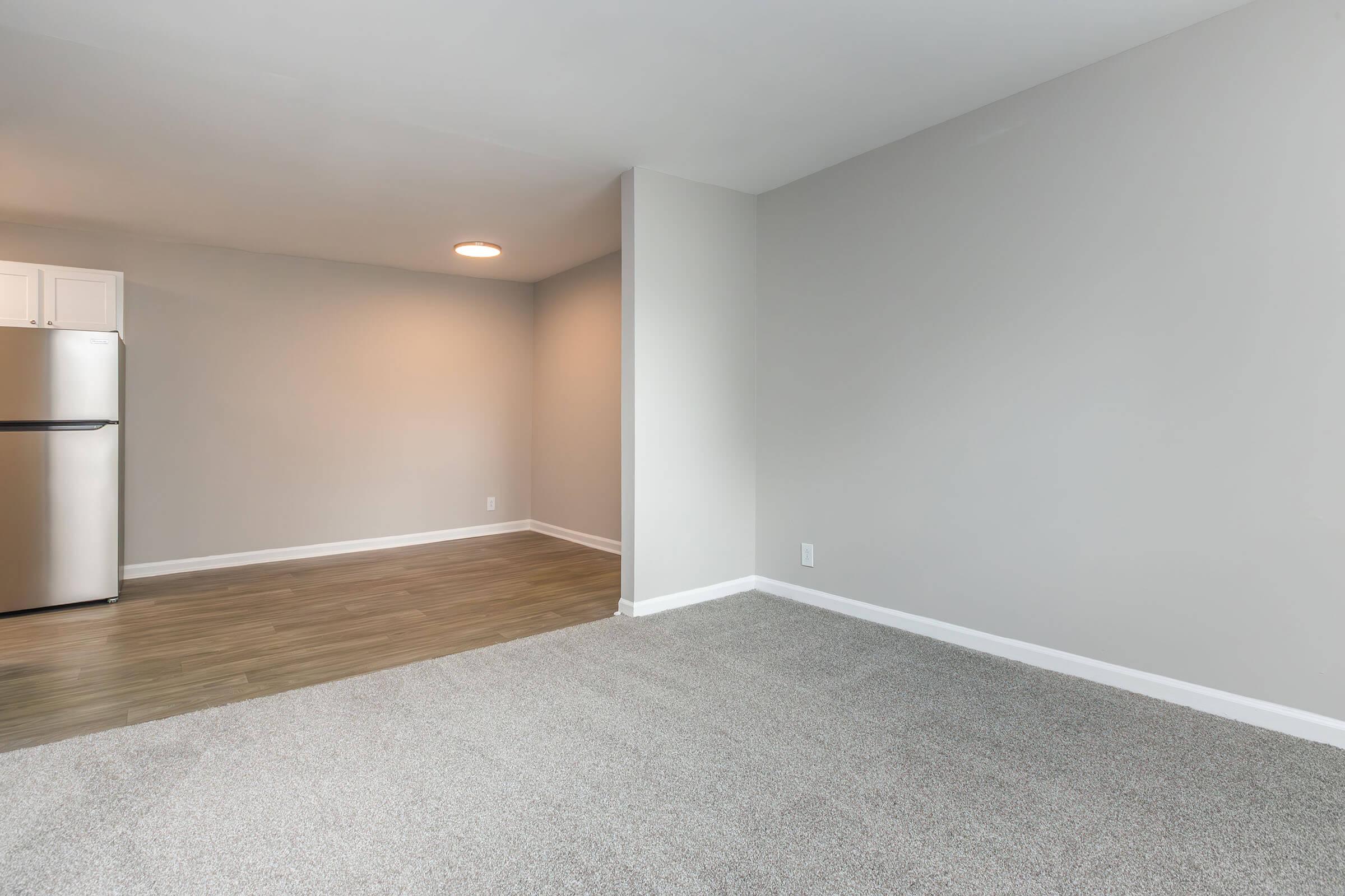 A spacious, empty room with light gray walls and new carpeting, featuring a corner leading to a kitchen area with a stainless steel refrigerator. Soft, natural light illuminates the space, enhancing the clean and modern feel.