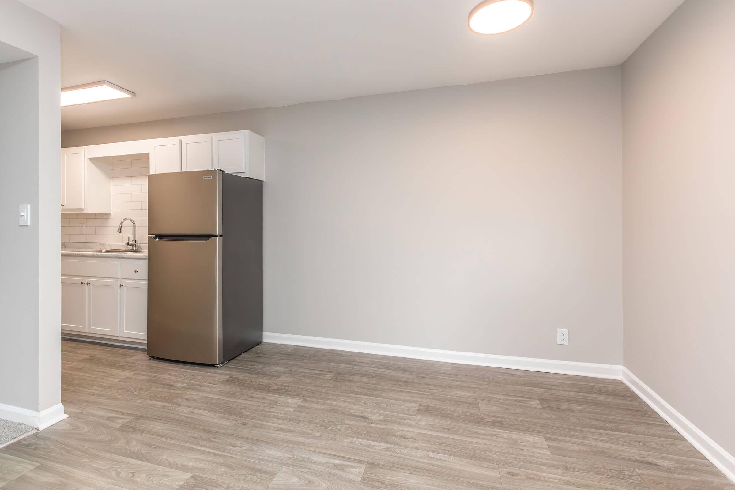 A modern, partially furnished kitchen and living area featuring a stainless steel refrigerator, white cabinetry, and light wood-like flooring. The walls are painted in a soft gray, and there is a ceiling light illuminating the space, creating a bright and open atmosphere.