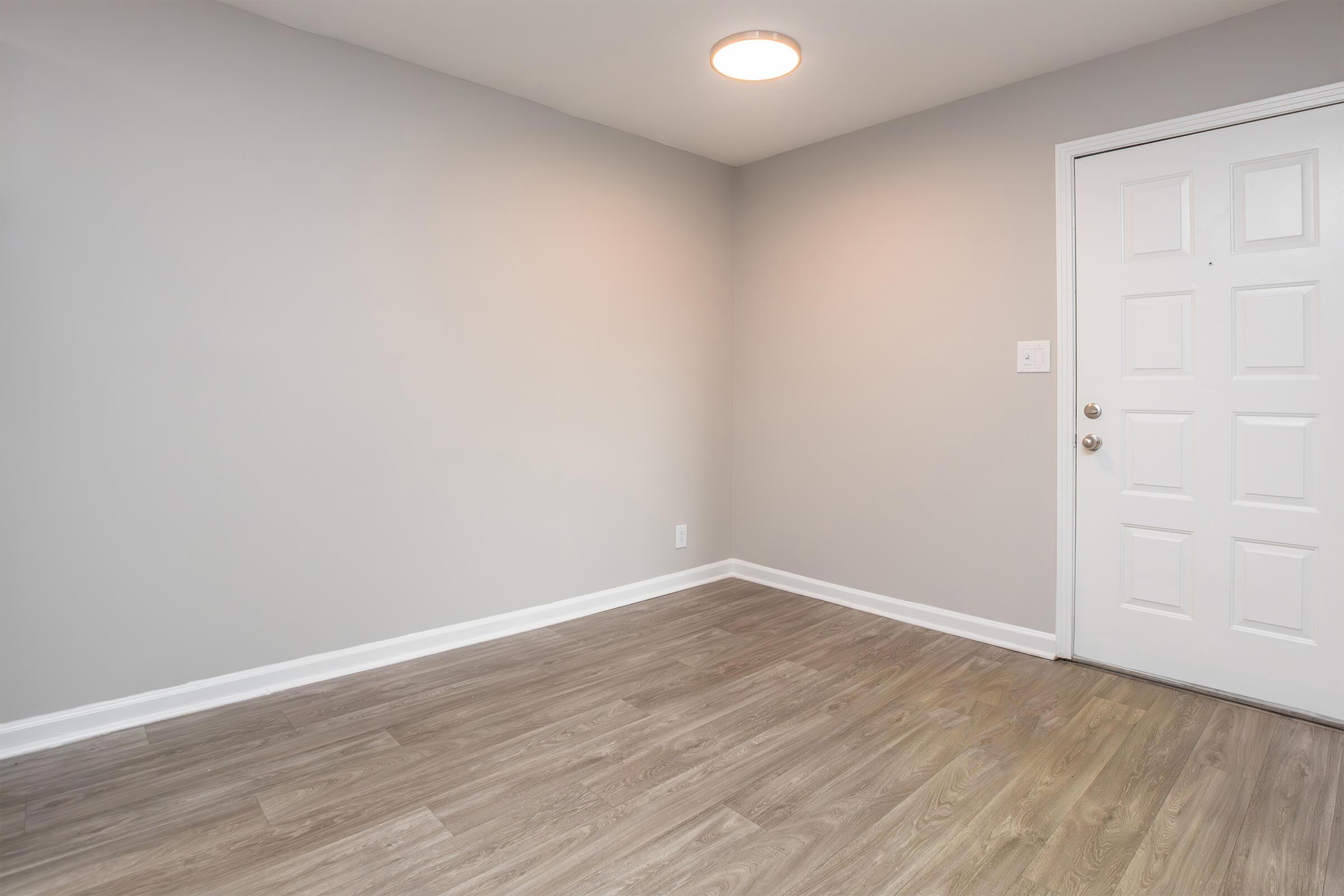 Empty room with light gray walls and laminate flooring. A white door with a doorknob is on the right side, and there is a ceiling light fixture. The space is uncluttered and provides a blank canvas for potential furnishing or decoration.