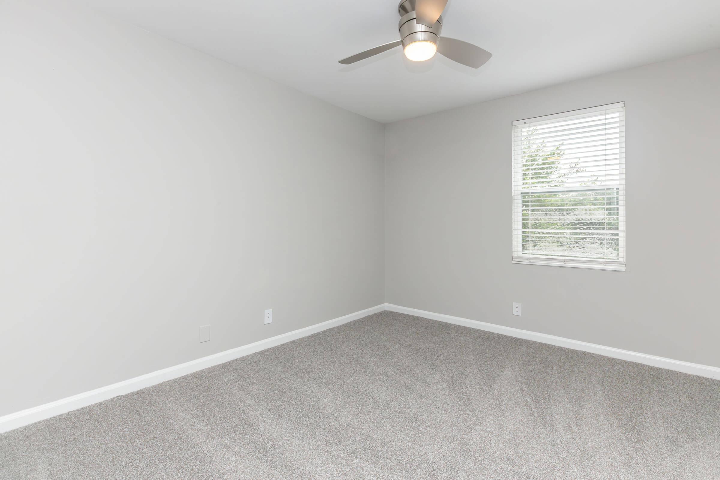A small, empty bedroom featuring light gray walls and a large window with blinds. The floor is covered in plush, light gray carpet. A modern ceiling fan is installed in the corner. The room has a simple and clean appearance, ideal for personalization and decoration.