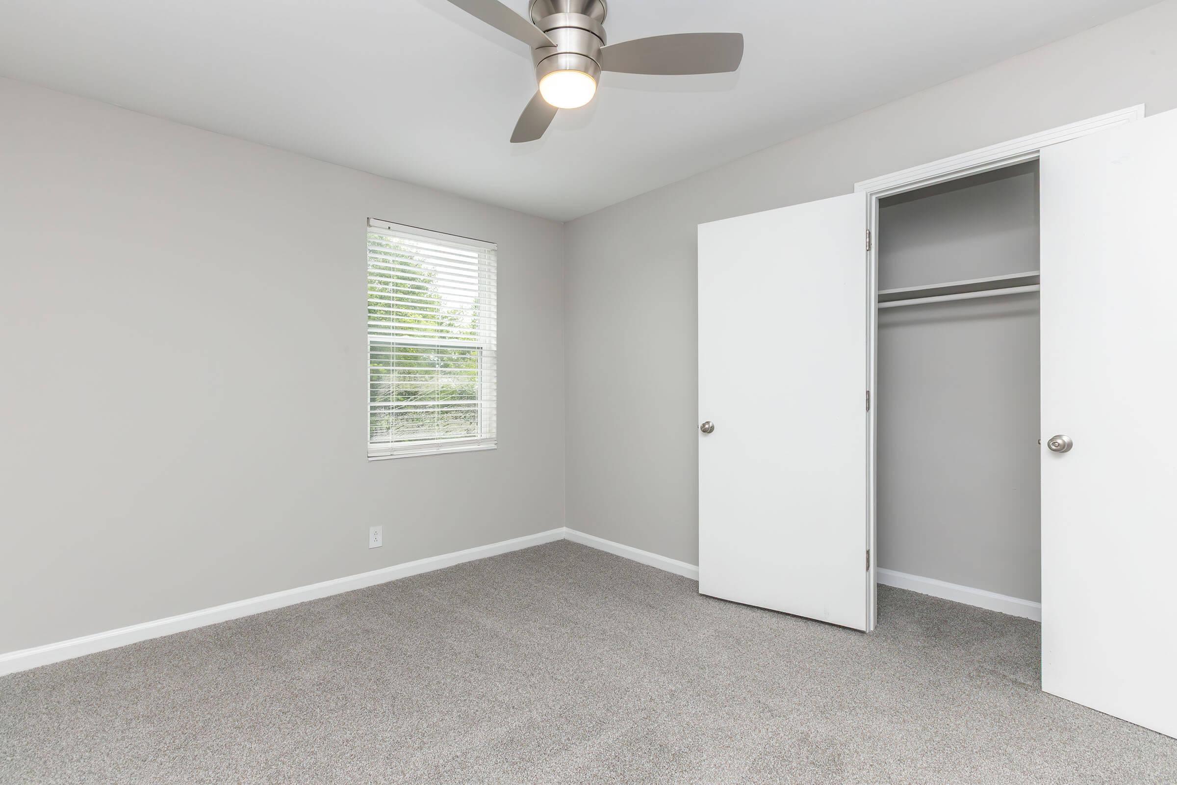 Empty room with light gray walls and carpeted floor. A white ceiling fan is installed, and there is a window with blinds allowing natural light in. An open closet with double white doors is visible on the right side of the image.