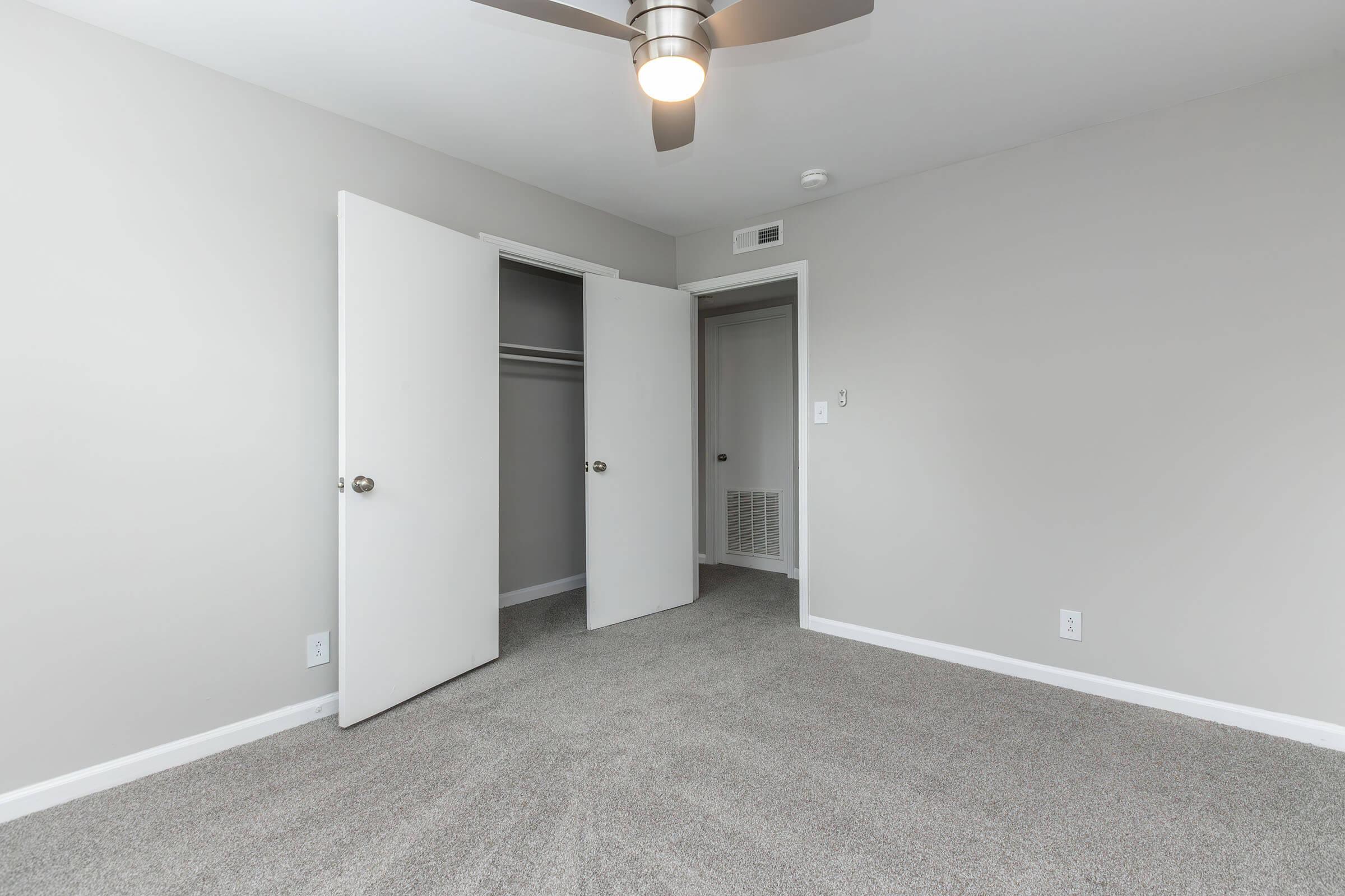 A neutral-colored bedroom featuring light gray walls and carpet. Two white doors are slightly open, revealing an empty closet space. A ceiling fan is installed, providing a modern touch. The room is bright and uncluttered, ideal for personalizing as a living space.