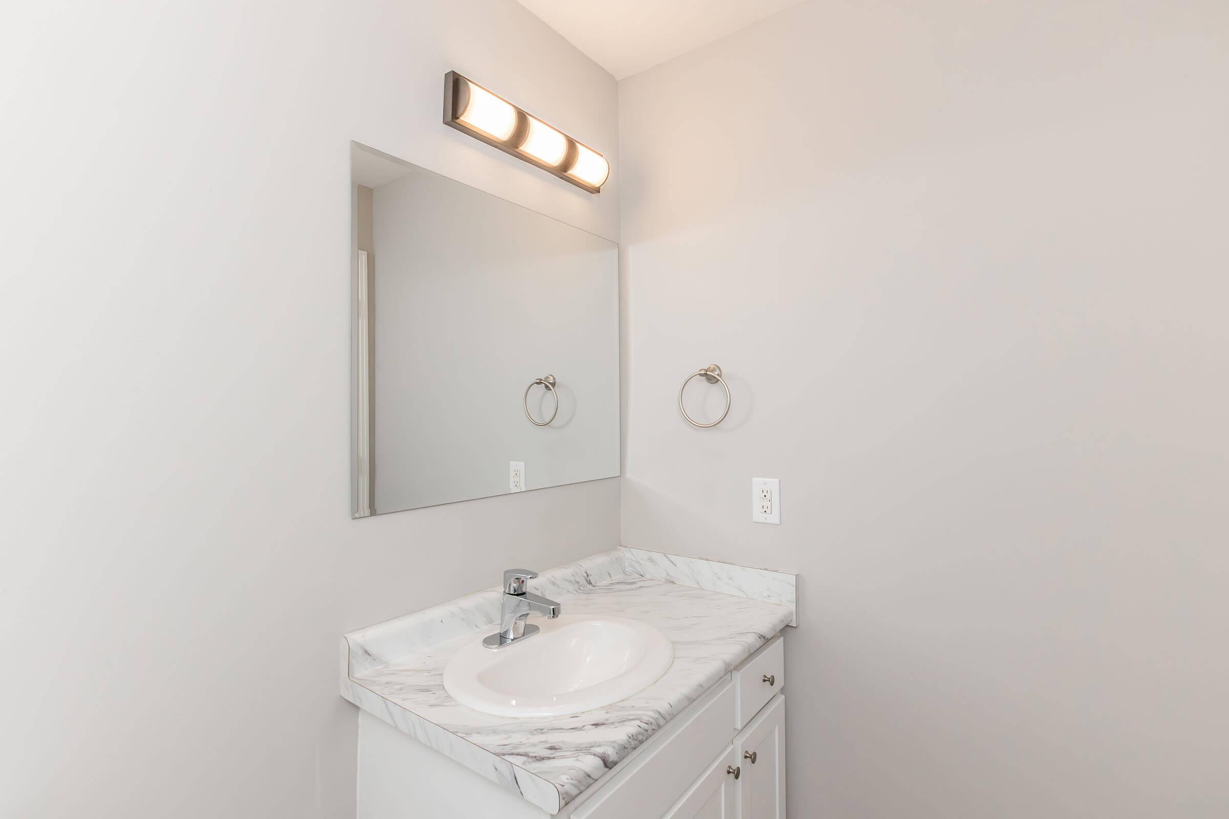 A modern bathroom with a light gray wall, a white marble countertop, and a round sink. Above the sink, there’s a rectangular mirror and a light fixture with three bulbs. A towel ring is mounted on the wall beside the sink. The overall design is clean and minimalist.