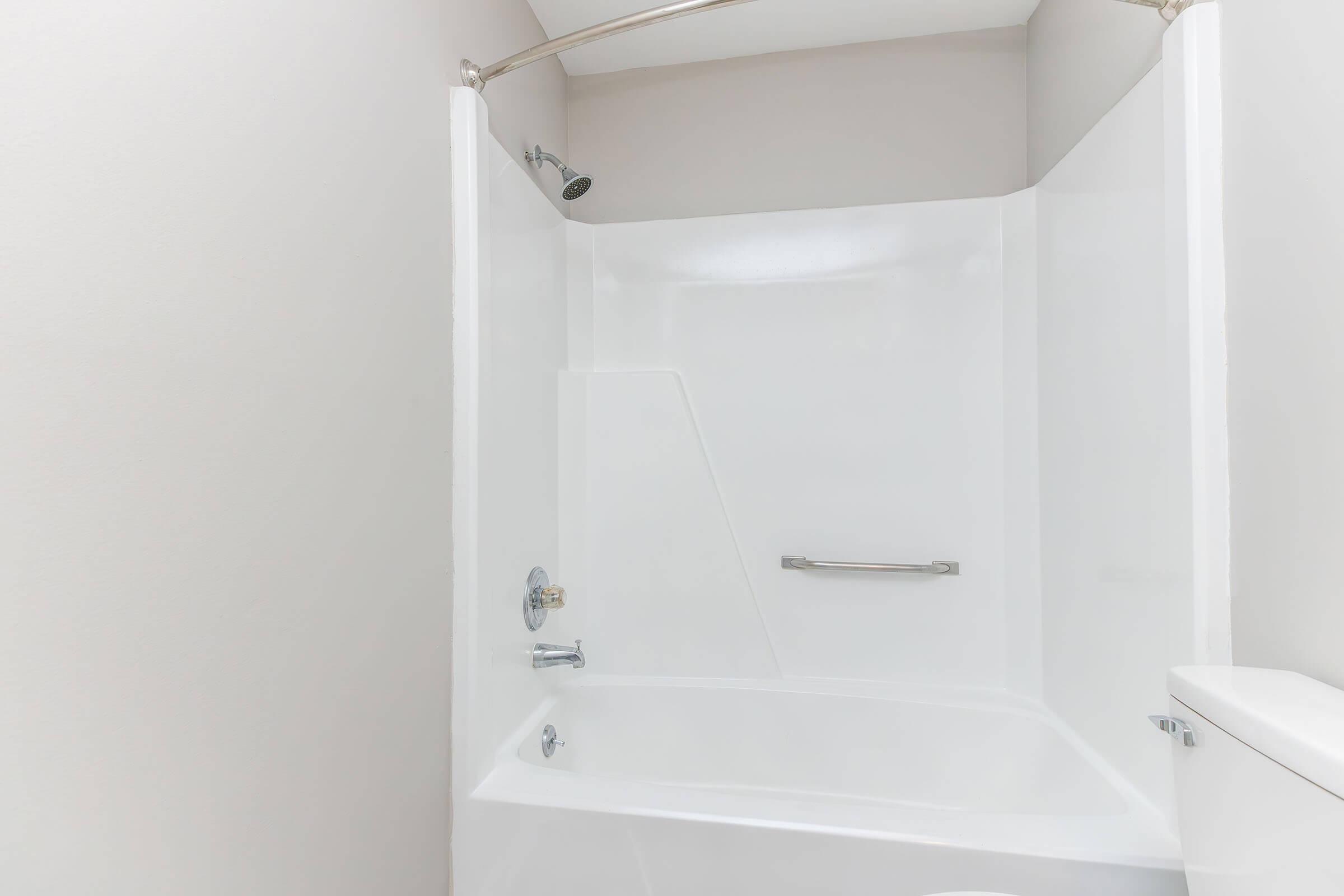 A clean, white shower stall with a shower head, a grab bar on the wall, and a bathtub section. The walls are plain and light-colored, creating a bright and modern appearance. The space is uncluttered and ready for use.