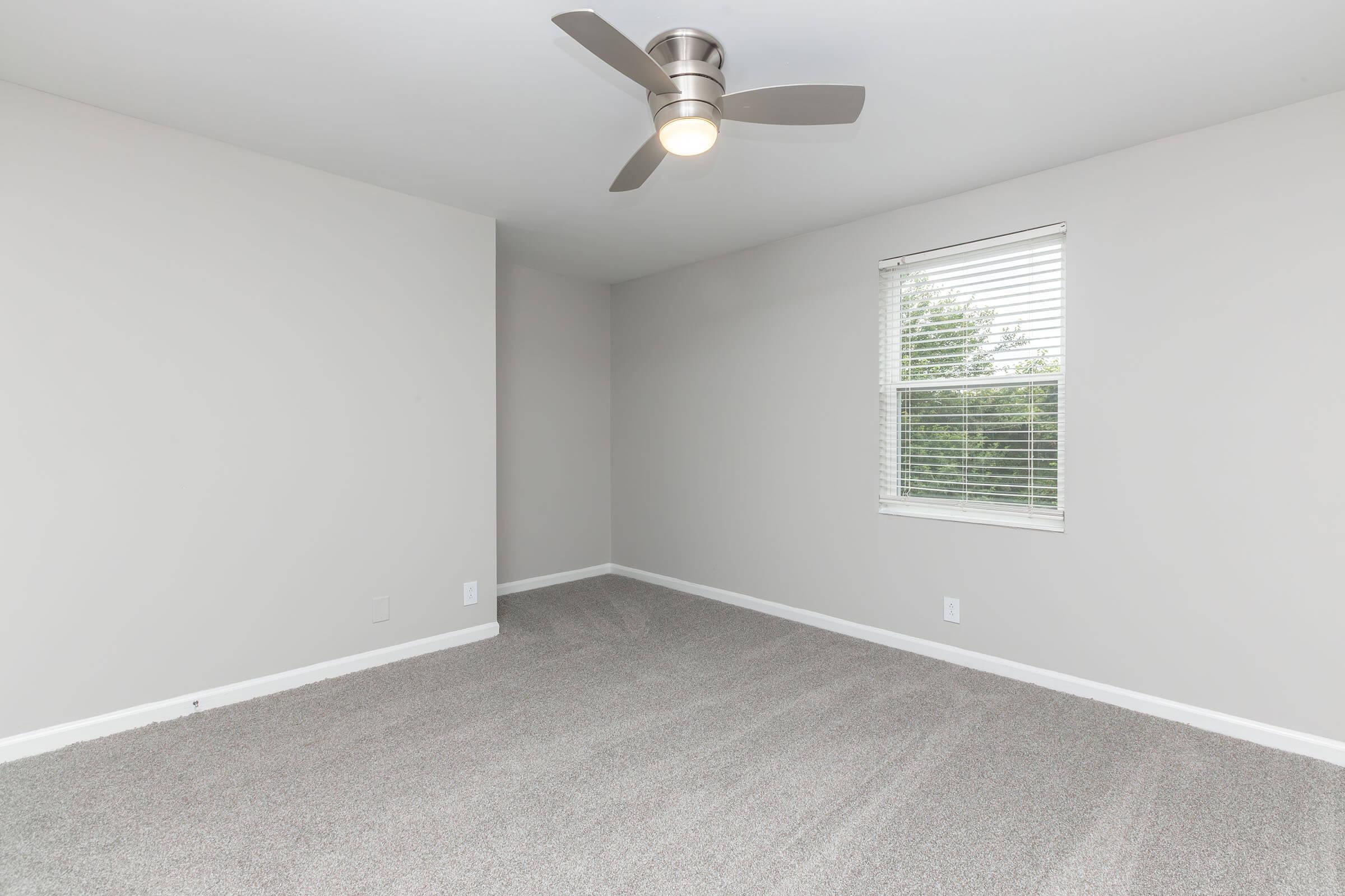 A spacious, empty room with light gray walls and new carpet. It features a modern ceiling fan and a window with white blinds that allows natural light to enter. The room has a clean and minimalist aesthetic, ideal for personalization or decorating.
