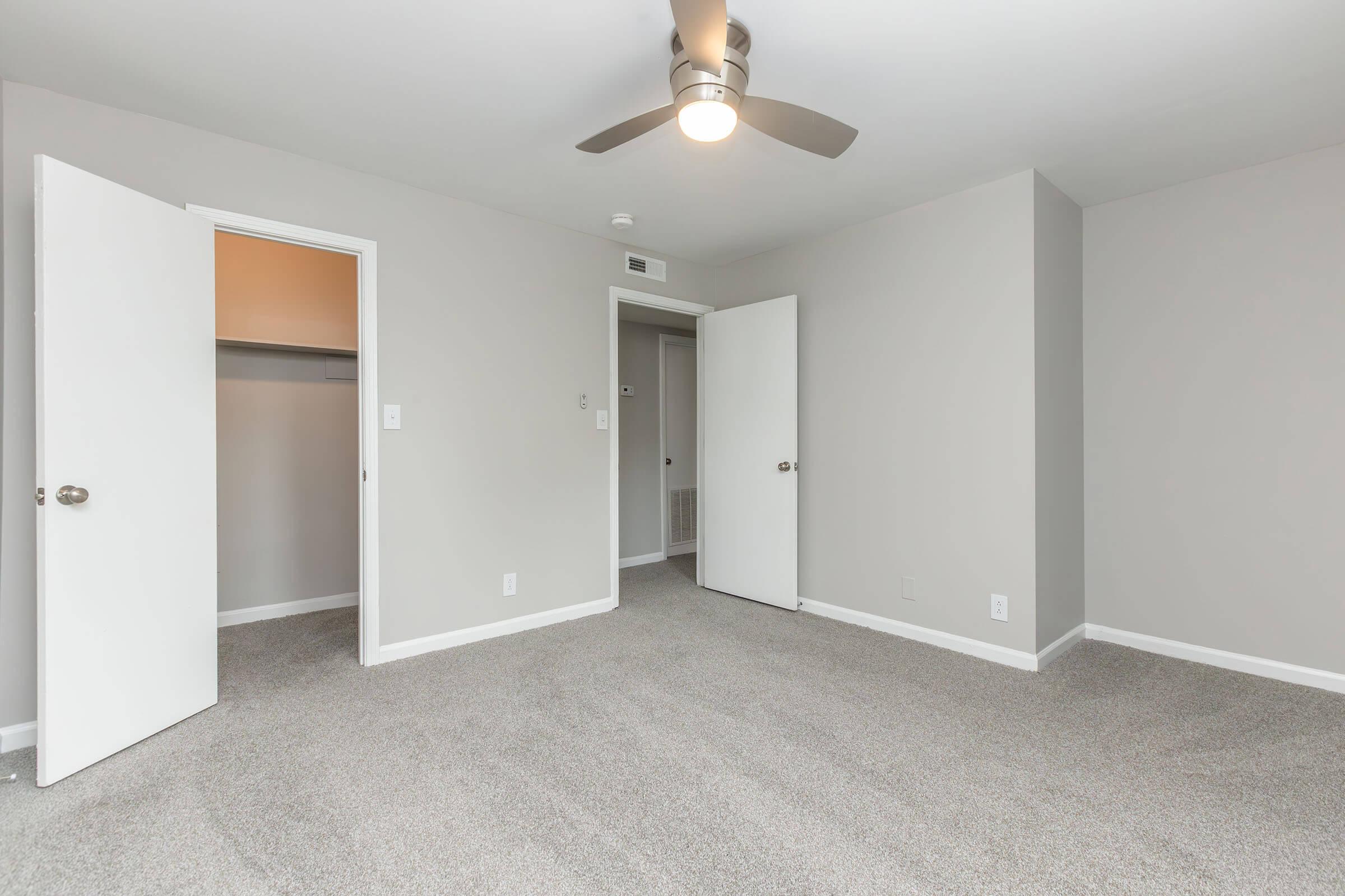 A spacious, empty room with light gray walls and carpeted flooring. Two white doors are open, revealing a closet and an adjacent room. A ceiling fan is installed in the center of the room, providing a modern touch to the minimalistic design. Natural light brightens the space through the open doors.