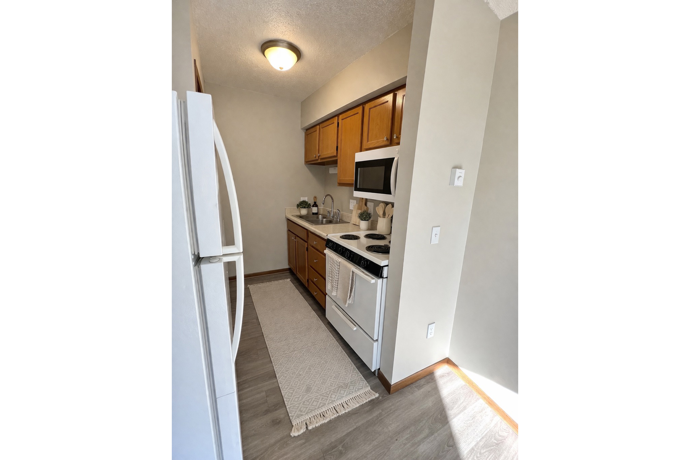 A small kitchen featuring light brown wooden cabinets, a white refrigerator, an electric stove, and an oven. The countertop is equipped with a microwave and a few kitchen utensils. A cream-colored rug lies on the floor, and light enters from a nearby window, creating a bright and inviting atmosphere.