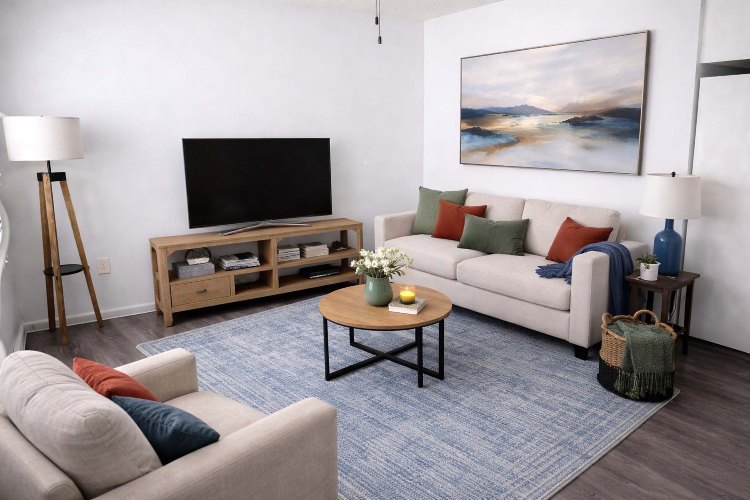 A cozy living room featuring a light-colored sofa with decorative pillows, a wooden coffee table, and an area rug. A large television is set on a wooden entertainment unit, while a piece of landscape artwork hangs on the wall. There are a lamp and a blue vase on the side, enhancing the inviting atmosphere.