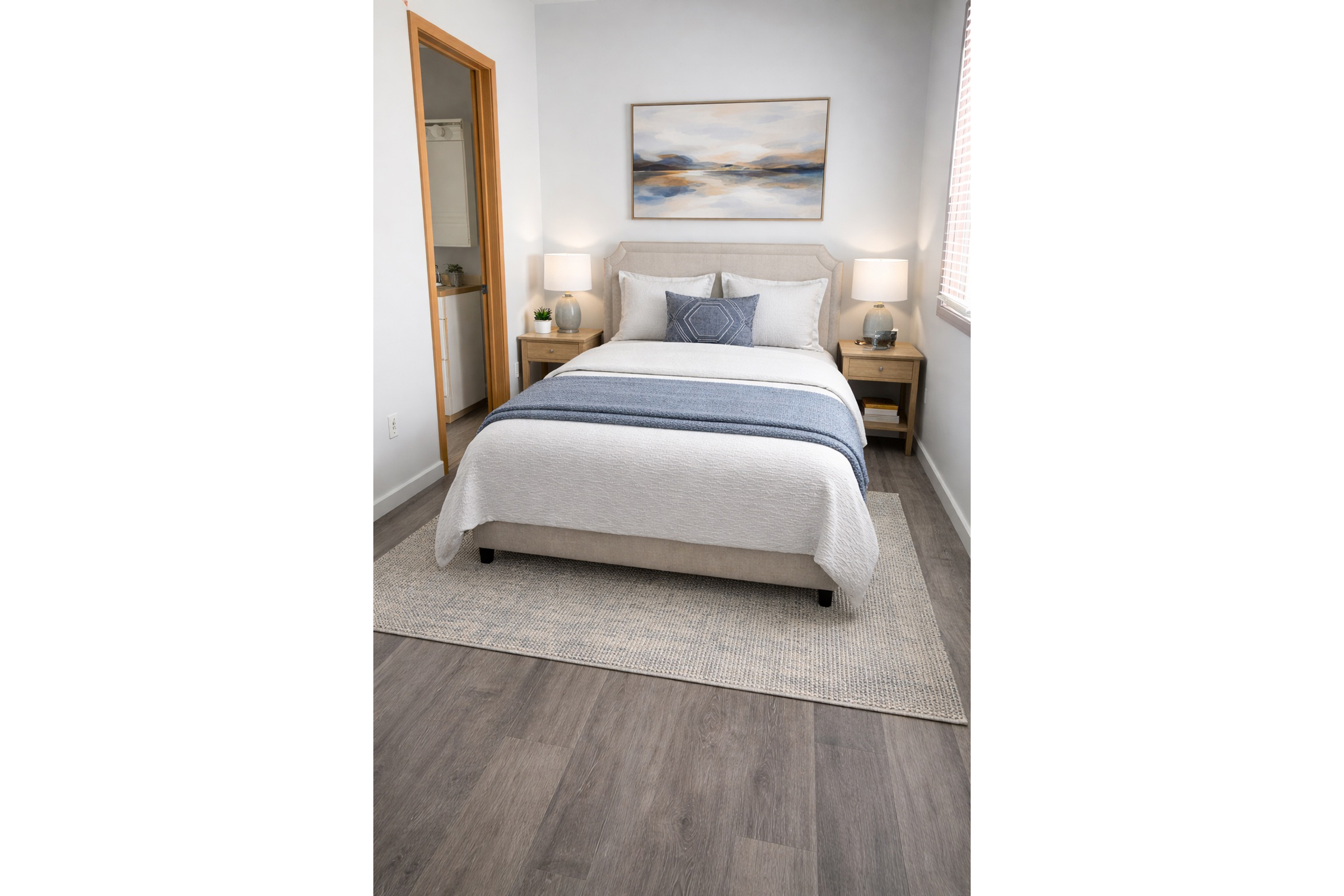 A modern bedroom featuring a queen-sized bed with a light gray comforter and blue accent stripes. There are two nightstands with lamps on either side, and a framed landscape painting above the bed. The walls are painted white, and light wooden flooring complements the décor. A door in the background leads to another room.