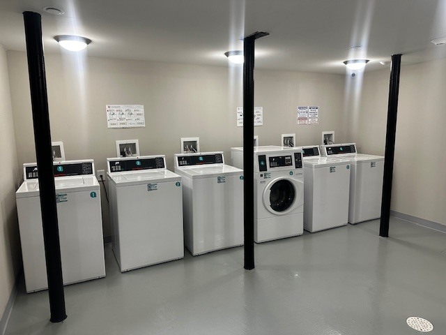 A laundry room featuring several white washing machines and a dryer lined up against a wall. The floor is gray, and the walls are light-colored. Overhead lighting is provided by fixtures on the ceiling, and there are instructional signs posted on the walls.
