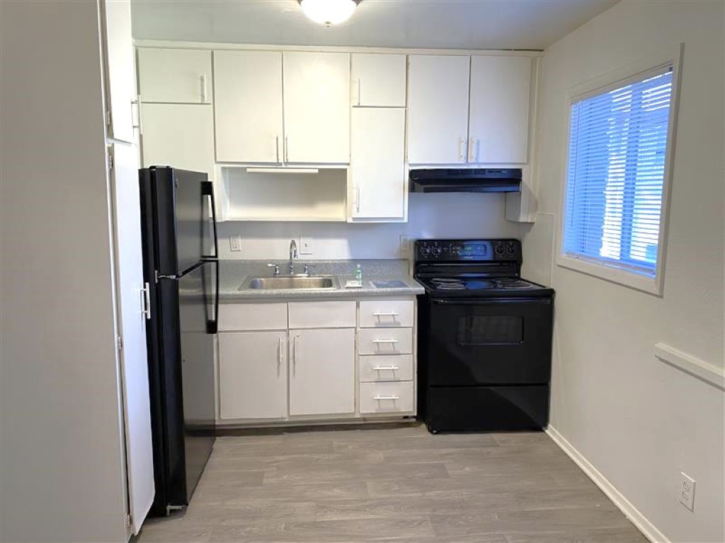 A compact kitchen featuring white cabinets, a black refrigerator, and a black stove. A stainless steel sink is located between them, with a window providing natural light. The kitchen has a light-colored floor and minimal decor, creating a clean and functional space.