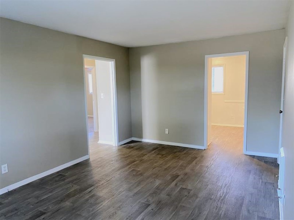 A spacious, empty room with light gray walls and a wooden floor. Two doorways on the left and right lead to other rooms, with natural light coming in from a window in the back. The overall atmosphere is clean and minimalistic.