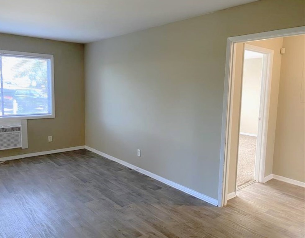 A vacant room with light brown walls and wood-like flooring, featuring a window with a view outside. An air conditioning unit is installed below the window. A doorway leads to another room on the right. The space is bright and uncluttered, ideal for various uses or decoration.