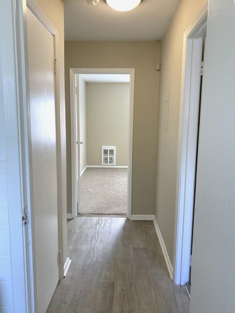 A view of a light-colored hallway featuring two white doors on the left, leading to separate rooms. The hallway has a wood-like floor and a neutral beige wall color. At the end of the hallway, a door leads to a carpeted room, which appears empty. A ceiling light illuminates the space.